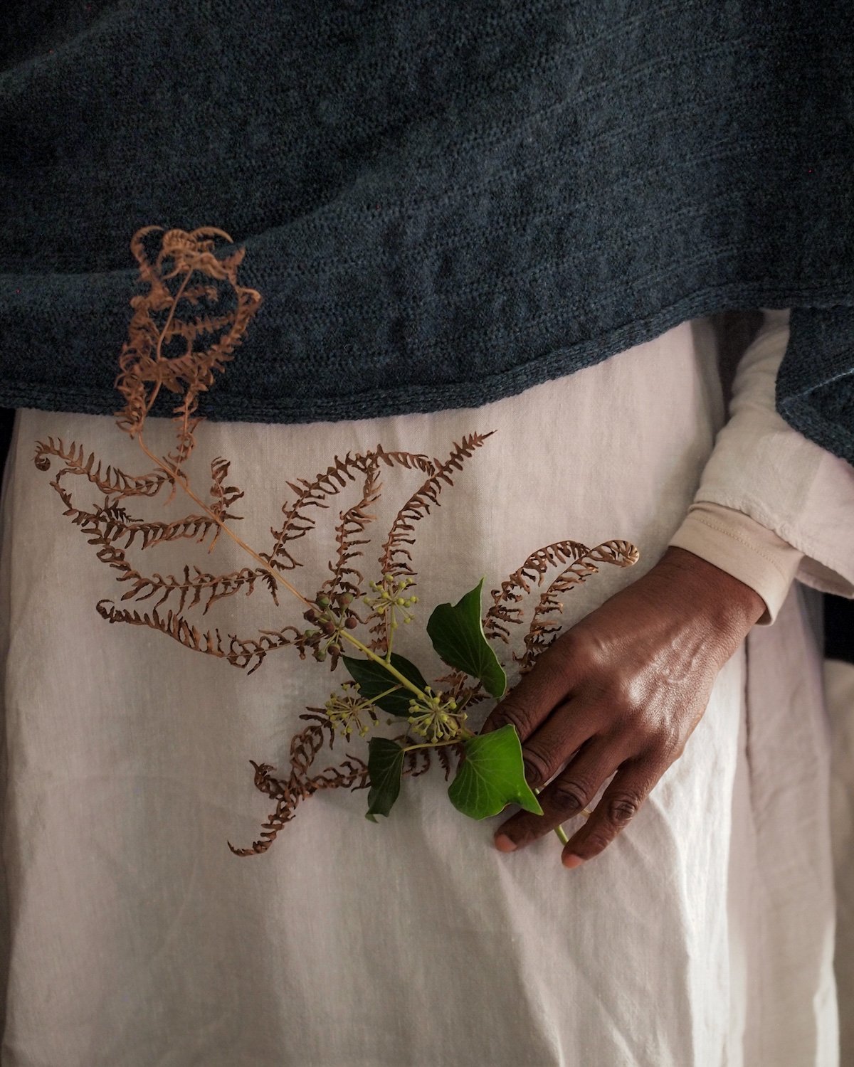 Person holding a bouquet of brown and green foliage, wearing a cream-colored long-sleeve shirt and a dark blue sweater.