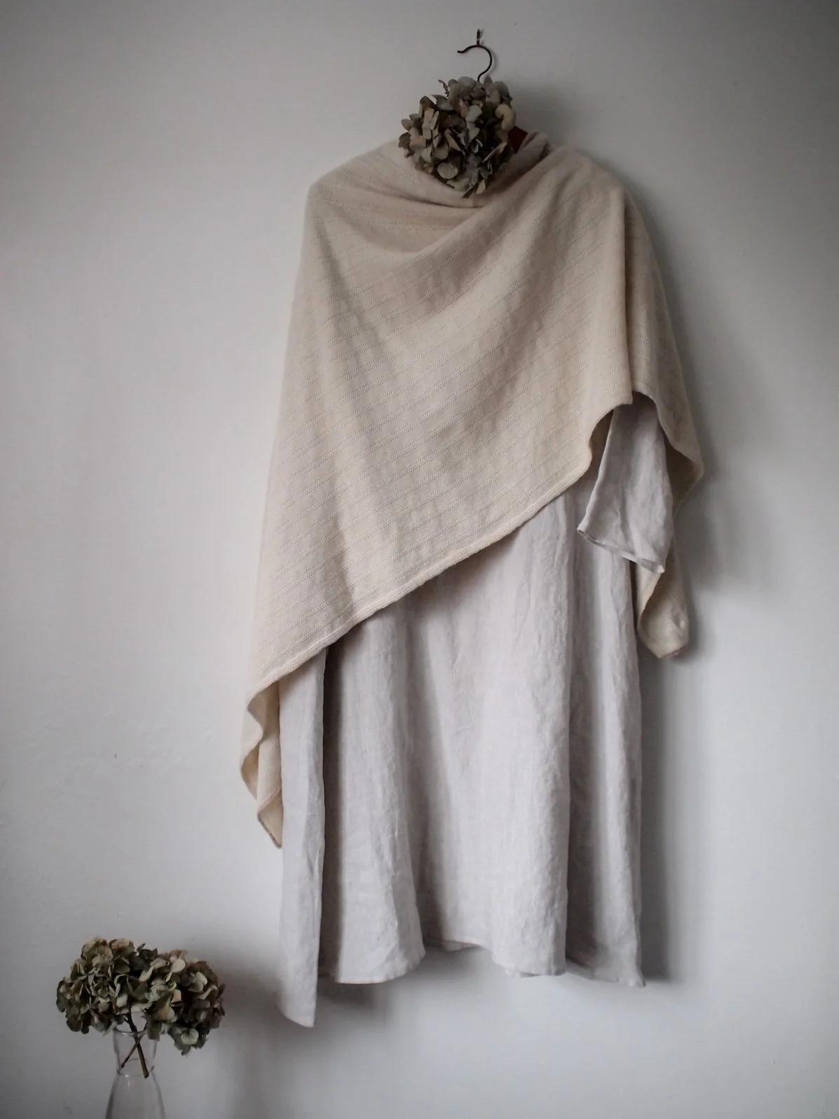 A cream-colored cloth hangs on a wall, with a dried flower arrangement attached at the top. A matching flower arrangement is in a glass vase on the floor below.