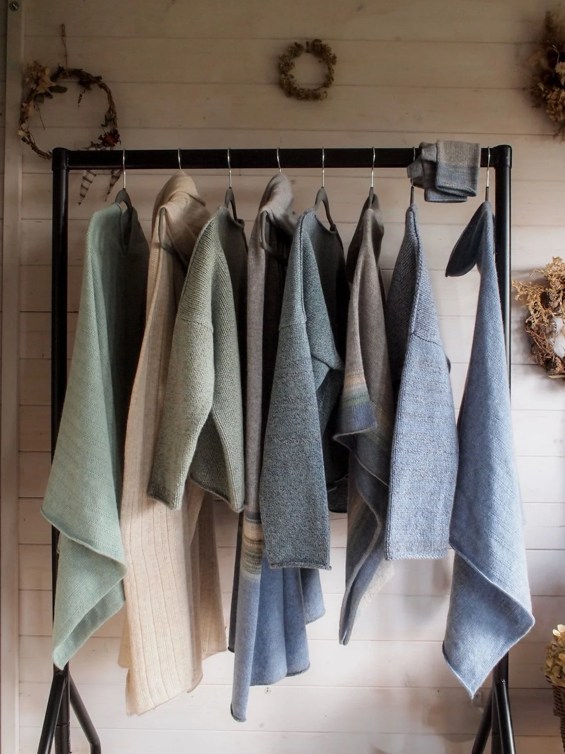A black clothing rack holding eight folded sweaters and seven hanging sweaters in neutral and muted tones, set against a light wooden wall with decorative wreaths.
