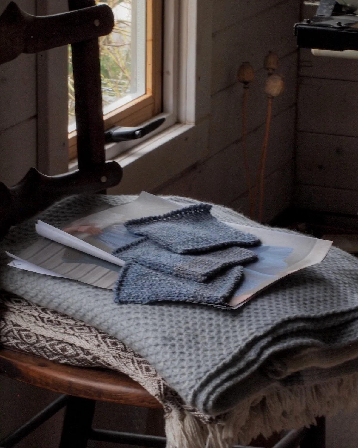 Knitted socks on top of fashion magazines and a patterned blanket, placed on a wooden chair near a window with a wooden wall in the background.