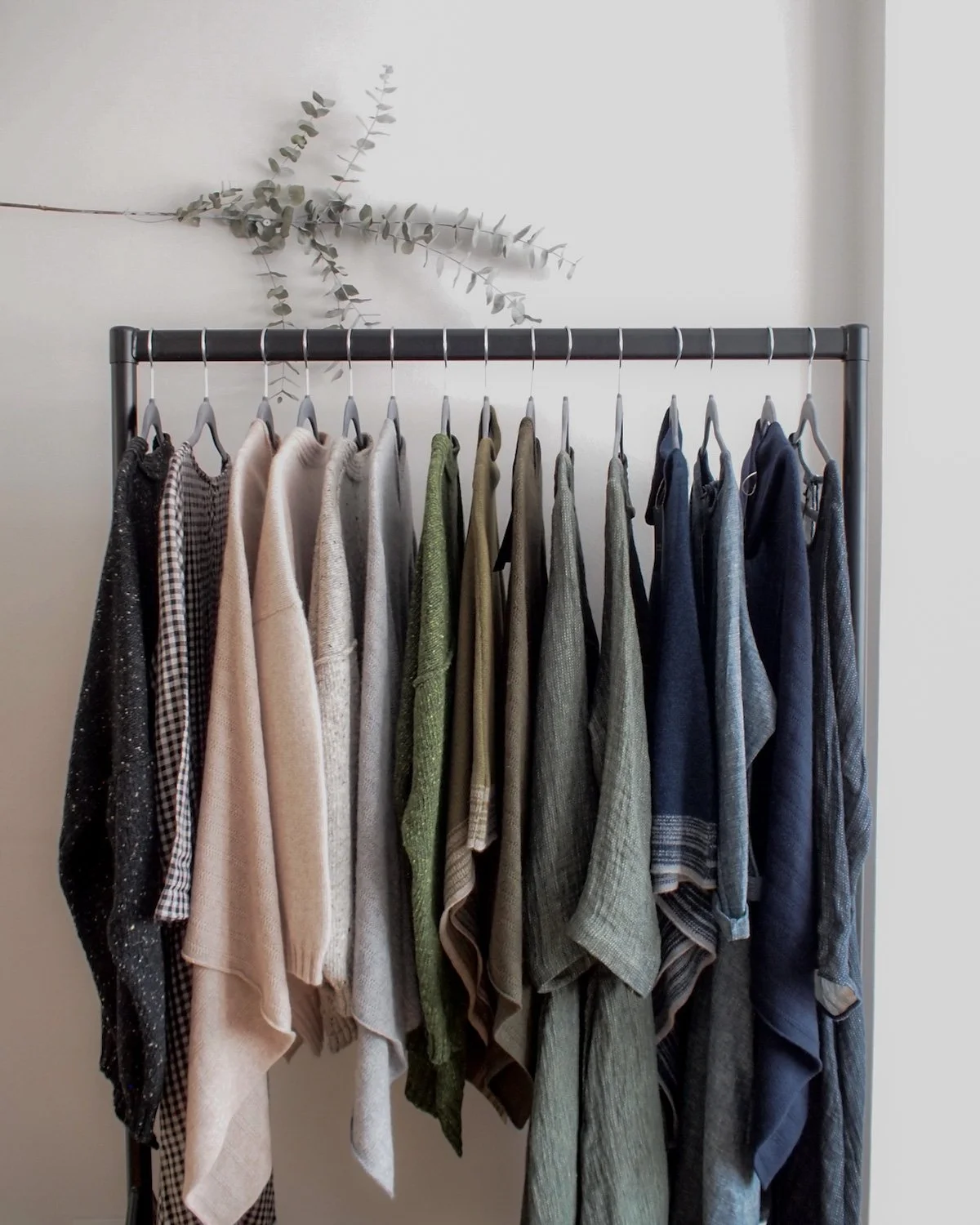 Clothes rack with various sweaters and shirts, olive branch decor above the rack, plain wall background.