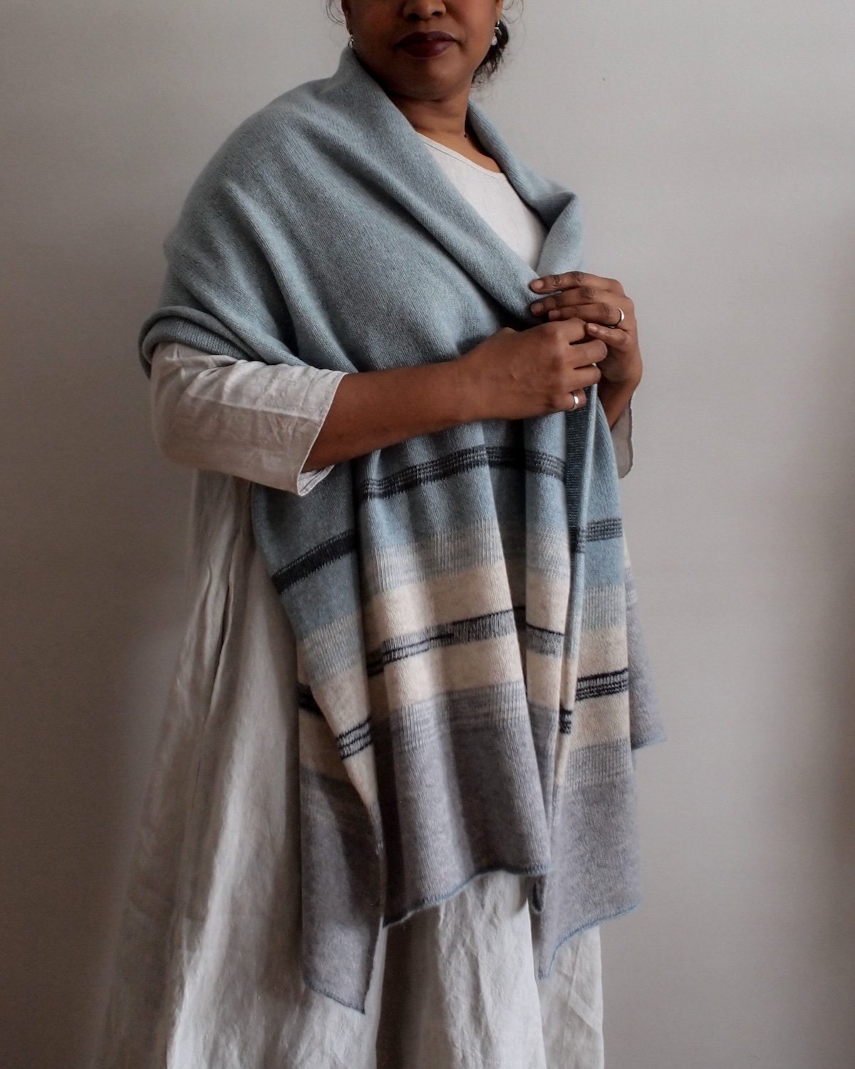 A woman wearing a beige dress with a white top underneath, draped in a gray and beige plaid shawl, standing against a plain light-colored wall.