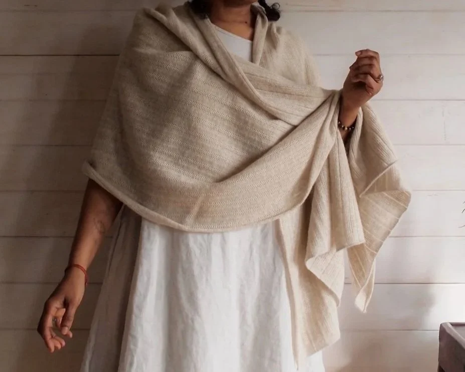 Person wearing a cream-colored shawl over a white dress, standing against a light wood-paneled wall.