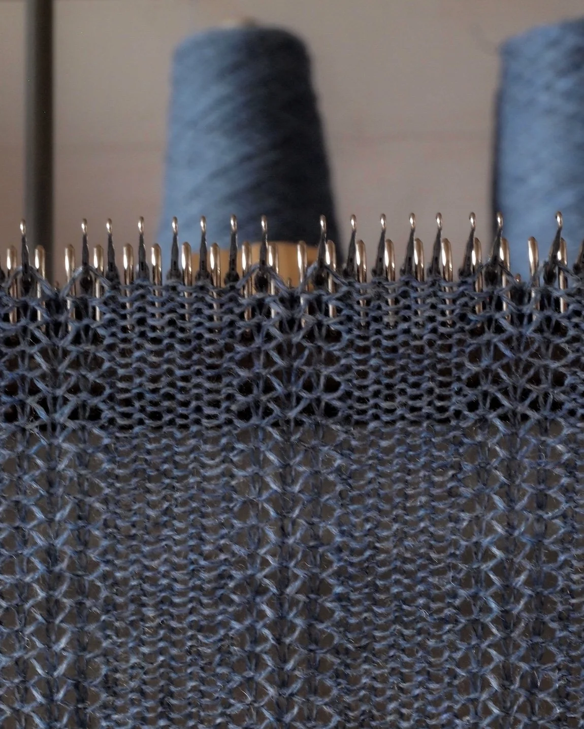 Close-up of a knitting project with metal knitting needles and gray yarn. Two spools of gray yarn are visible in the background.