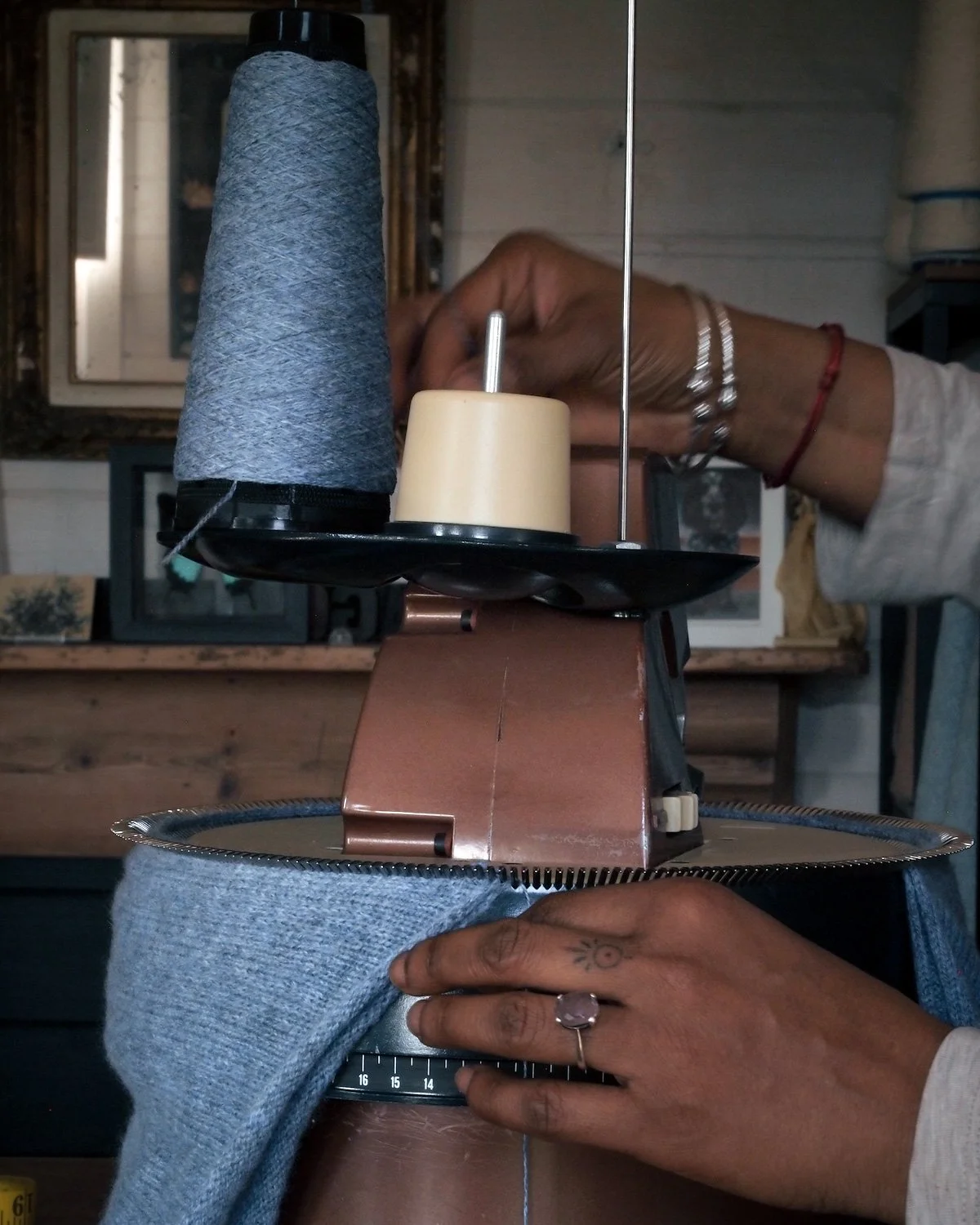 A person is sewing or weaving on a vintage sewing machine, with their hand placed on the metal part of the machine. The person wears bracelets, a ring, and has tattoos on their hand. The background includes picture frames and furniture.