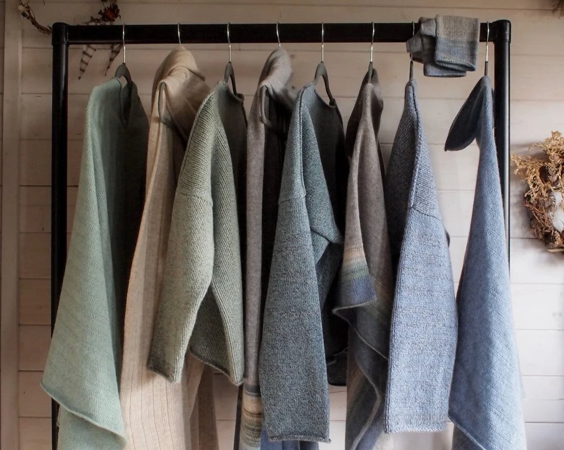 A clothing rack with nine sweater or jacket items hanging on hangers, displayed in various shades of gray and beige, against a wooden wall background.