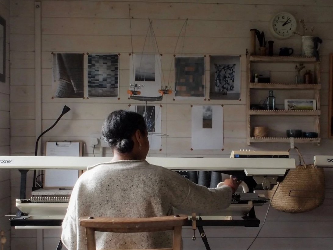 A woman sitting at a large embroidery or sewing machine in a cozy room with wooden walls. There are pictures, ornaments, and a clock on the wall next to shelves with various items.