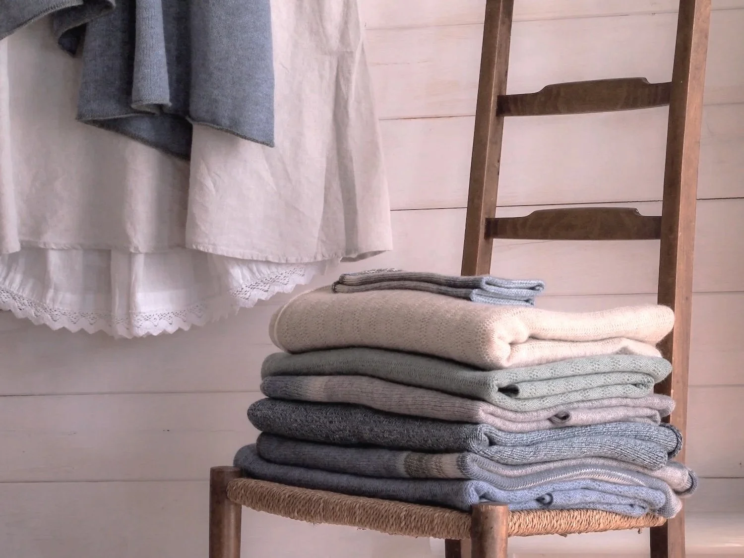 Stacked folded sweaters on a wooden chair next to hanging clothes against a light-colored wooden wall.