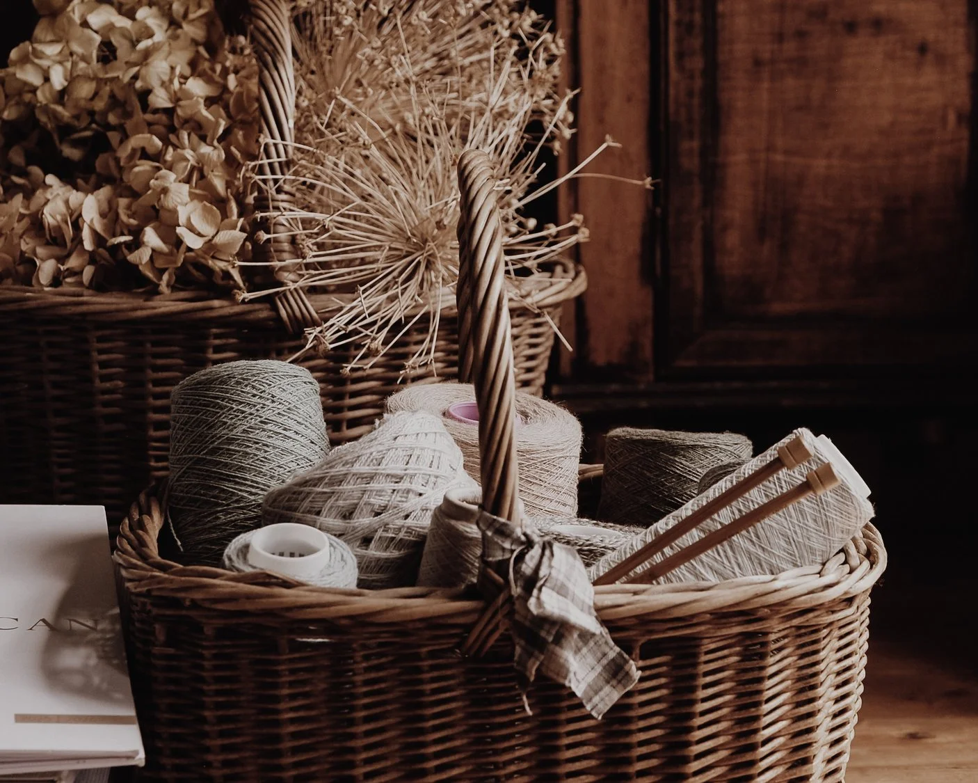 A wicker basket containing natural yarns and knitting needles. Another basket is behind with dried hydrangea and seed heads.