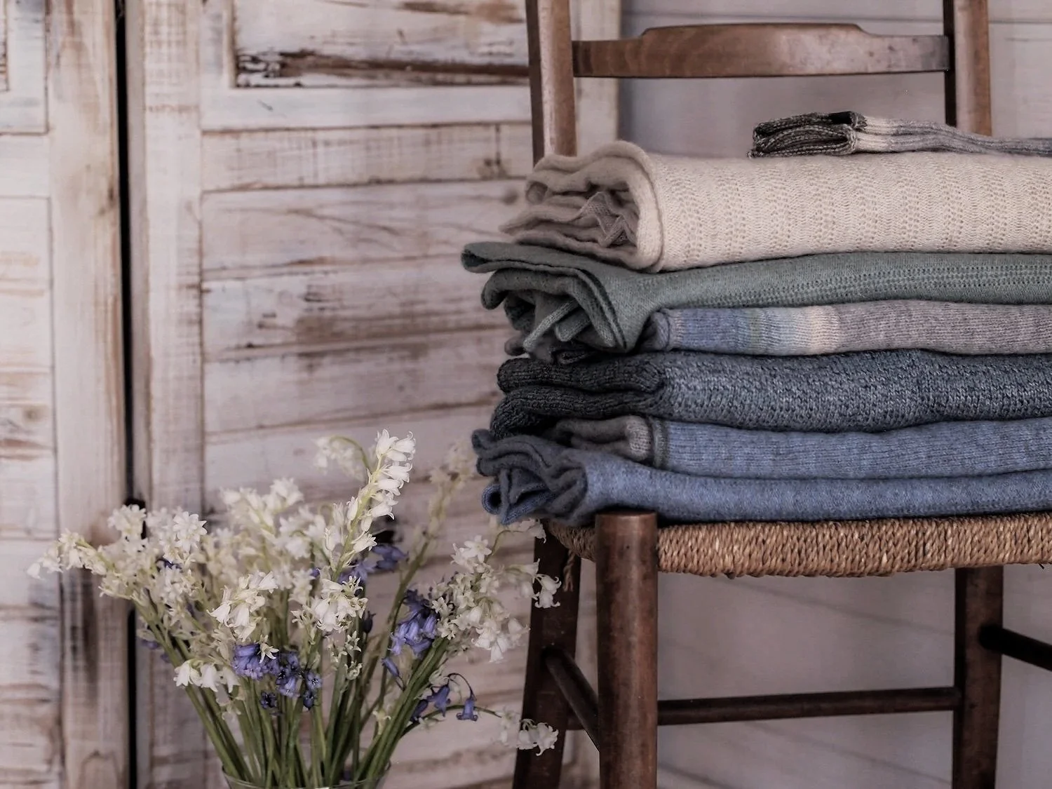 Stack of folded gray, beige, and blue sweaters on a wooden chair beside a vase of white and purple flowers against a white wooden wall.