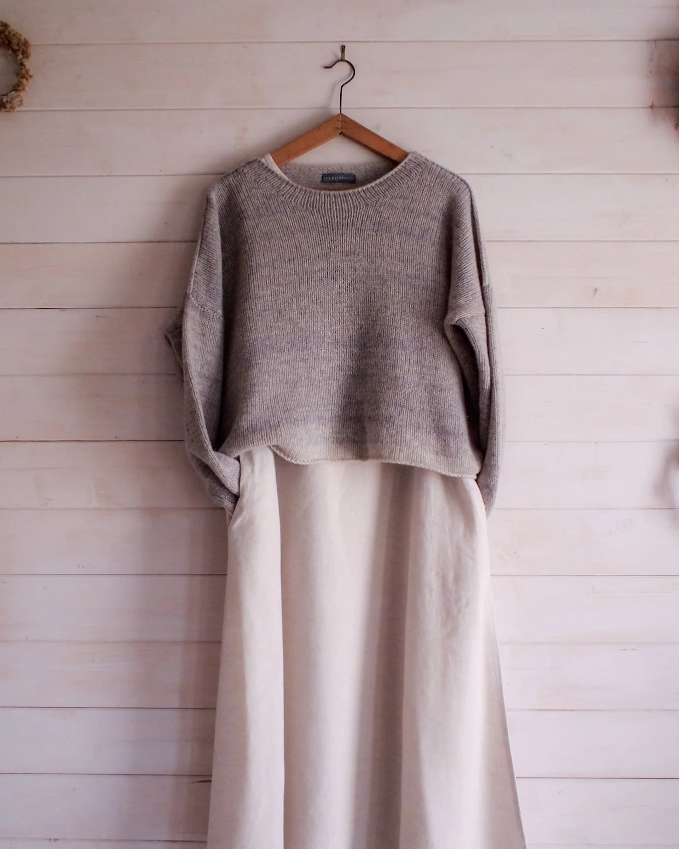 An ombré jumper hangs on a wall, with a dried flower arrangement attached at the top. A matching flower arrangement is in a glass vase on the floor below.