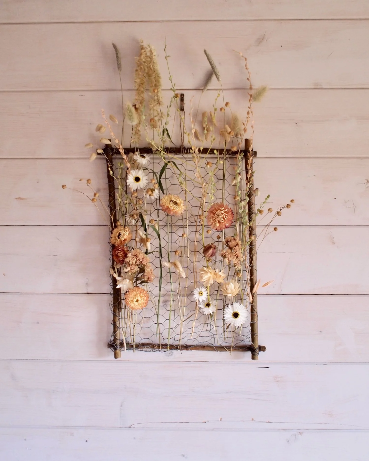 Willow frame with dried flowers woven through a chicken wire mesh Hung on the wall of Jules Hogan's white washed wooden walled studio.