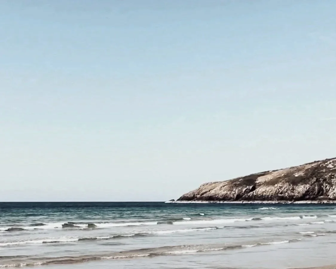 A calm beach with gentle waves and a rocky hillside in the background under a clear sky.