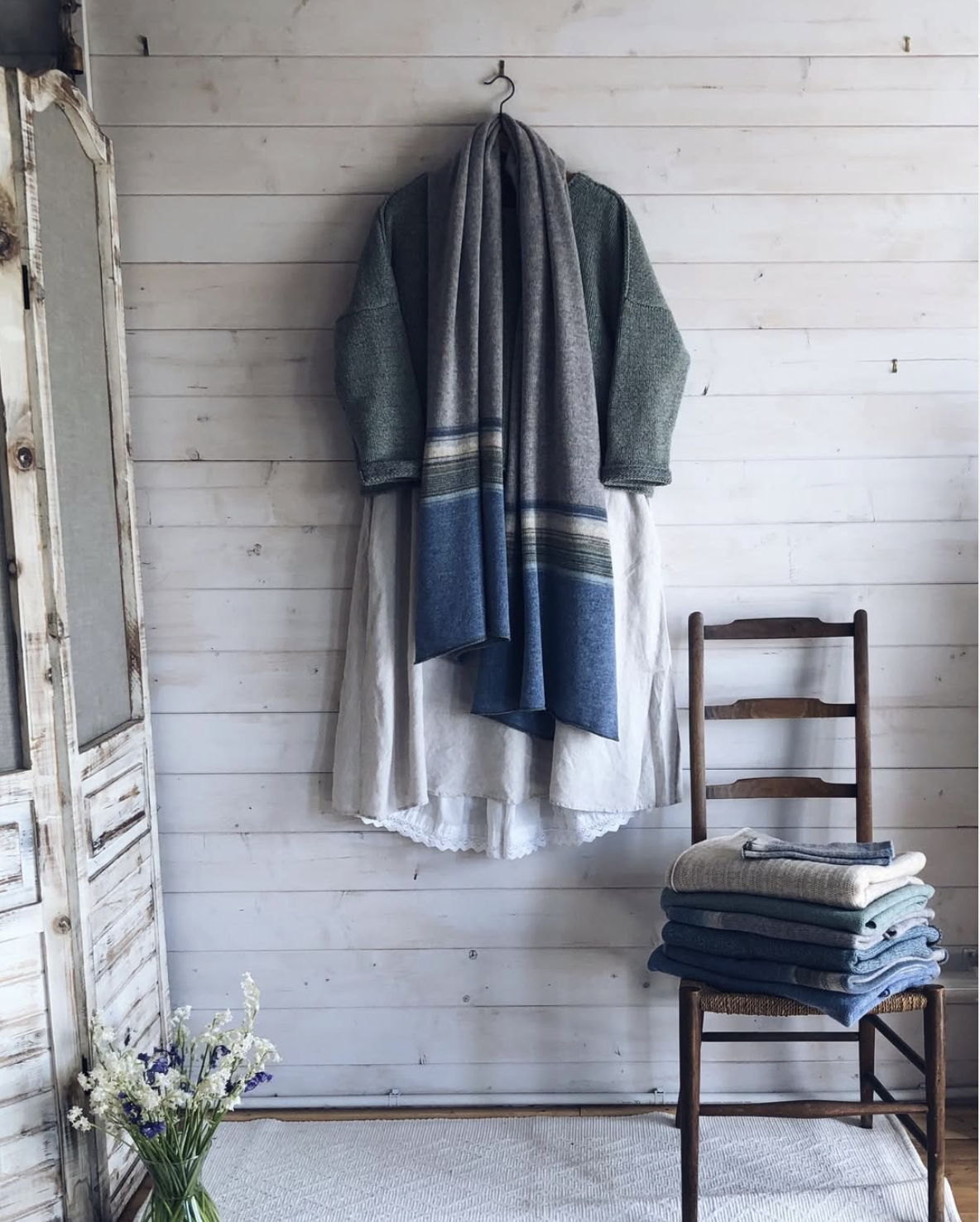 A white wooden wall with a coat hanging on a hook, a wooden chair with folded clothes on it, and a vase of flowers on a beige rug.