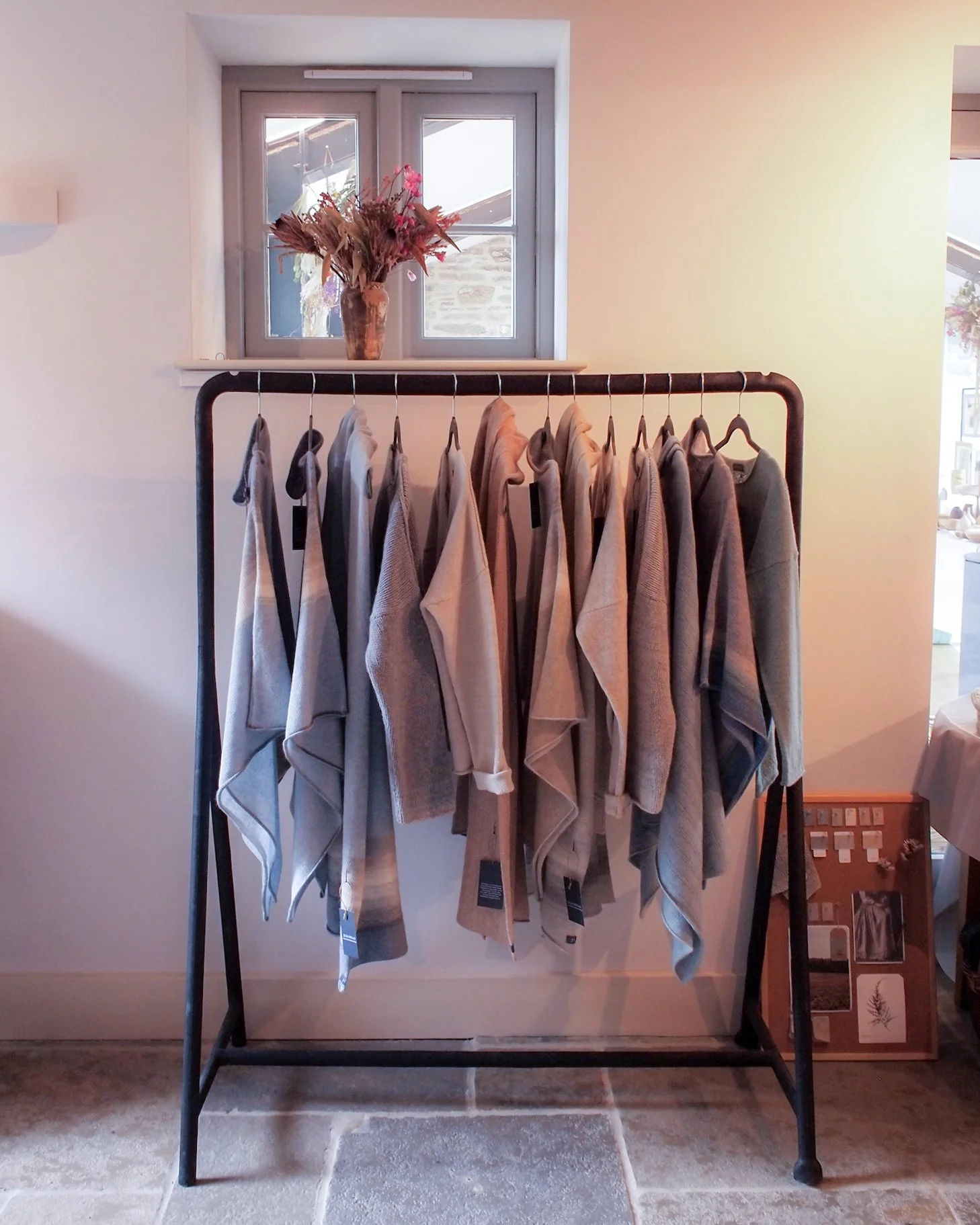 light airy room with a window at the back, a vase of dried flowers rests on the window sill. By the wall there is a rail of knitwear with soft neutral colour and blue/greys.