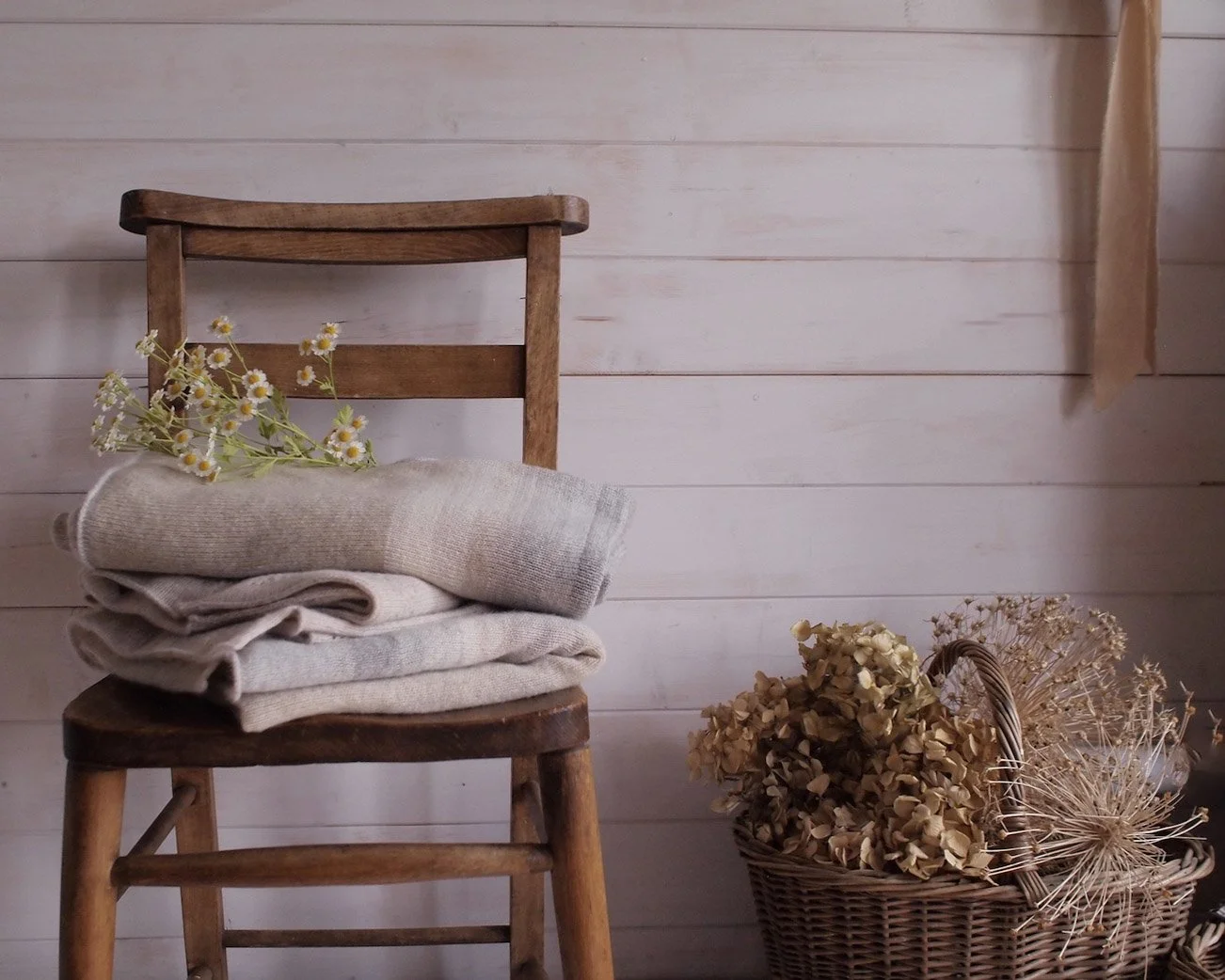 A wooden chapel chair with a pile of folded knits. Some fever few rests on the top. Beside the chair is a basket filled with hydrangea and large allium heads.