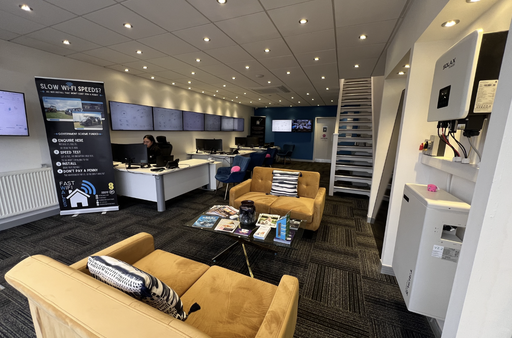 An indoor office waiting area with yellow sofas, a glass coffee table with magazines and a jar, and a row of desks with computer monitors and offices chairs in the background. A woman is sitting at one of the desks. A large black poster stands beside her advertising slow Wi-Fi speeds, along with a series of digital screens on the wall. To the right, there is a staircase, and a white solar inverter and a water dispenser on the wall.