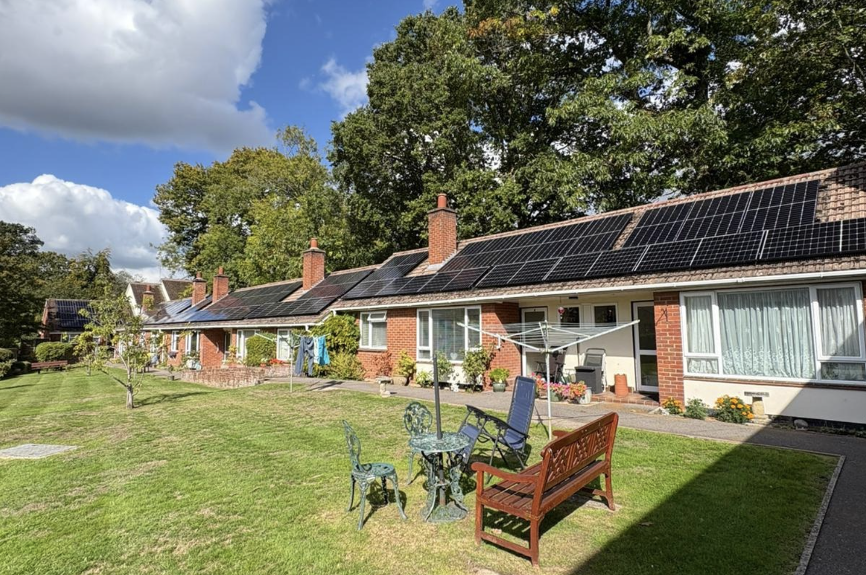 Backyard view of a row of houses with solar panels on the roofs, a well-maintained lawn, outdoor seating, a clothesline, and lush green trees under a partly cloudy sky.