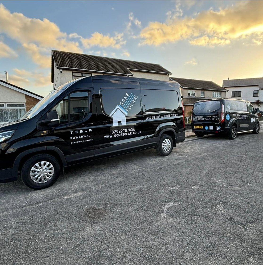 Two black vans parked on a residential street during sunset. The vans display solar energy company branding, including a house logo, website URL, and text about solar power installation.