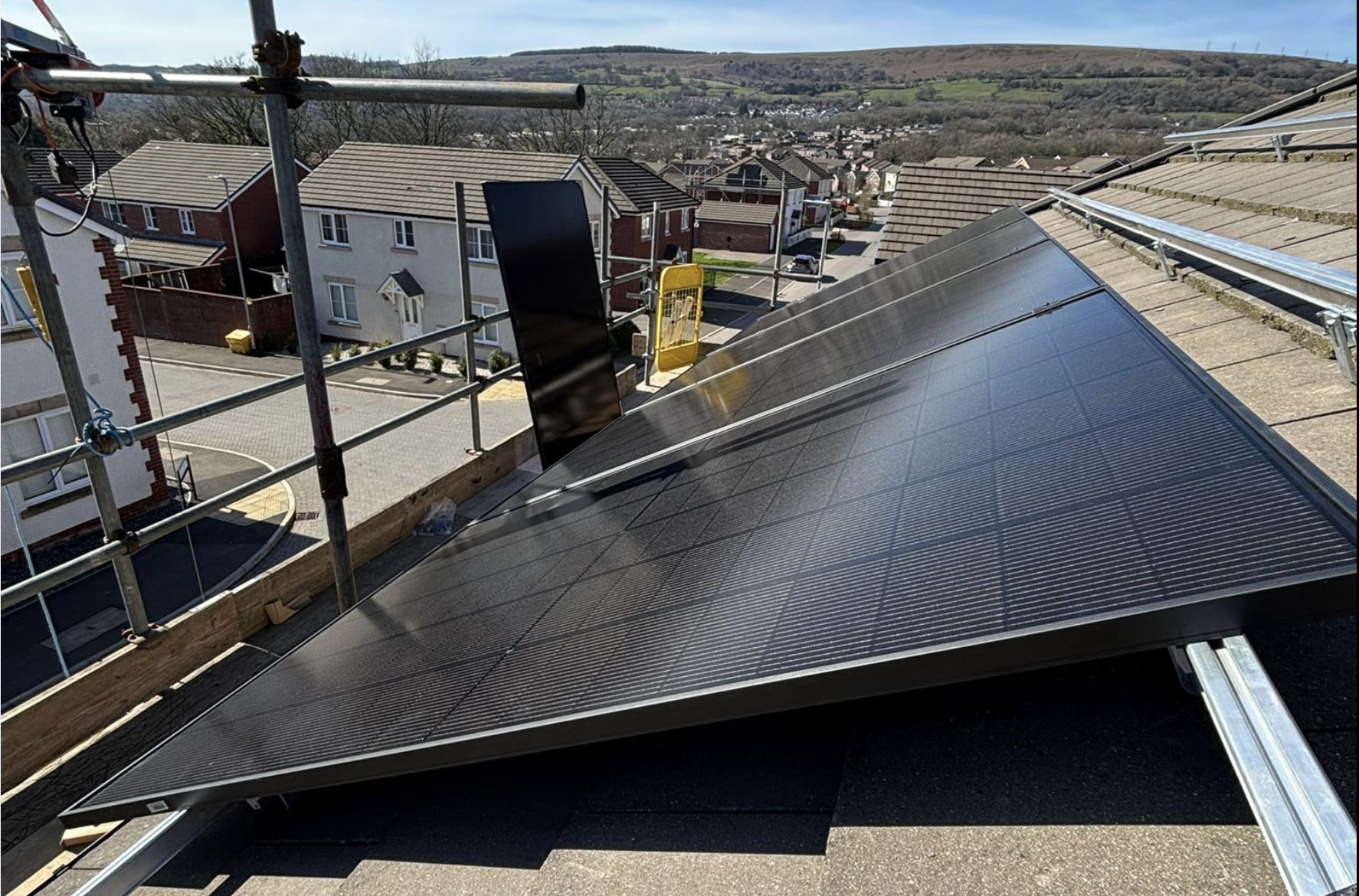 View of solar panels installed on the roof of a building under construction with scaffolding around, overlooking a residential area and hilly landscape in the distance.