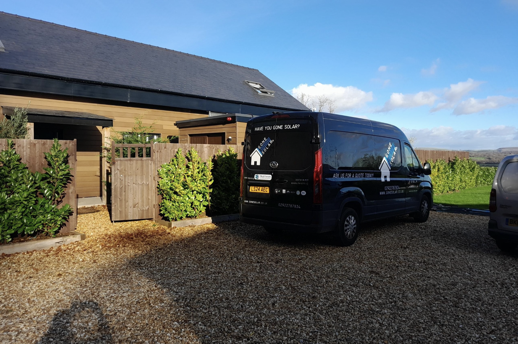 Black van with company branding parked in front of a modern house with wooden fencing and green bushes, under a partly cloudy blue sky.