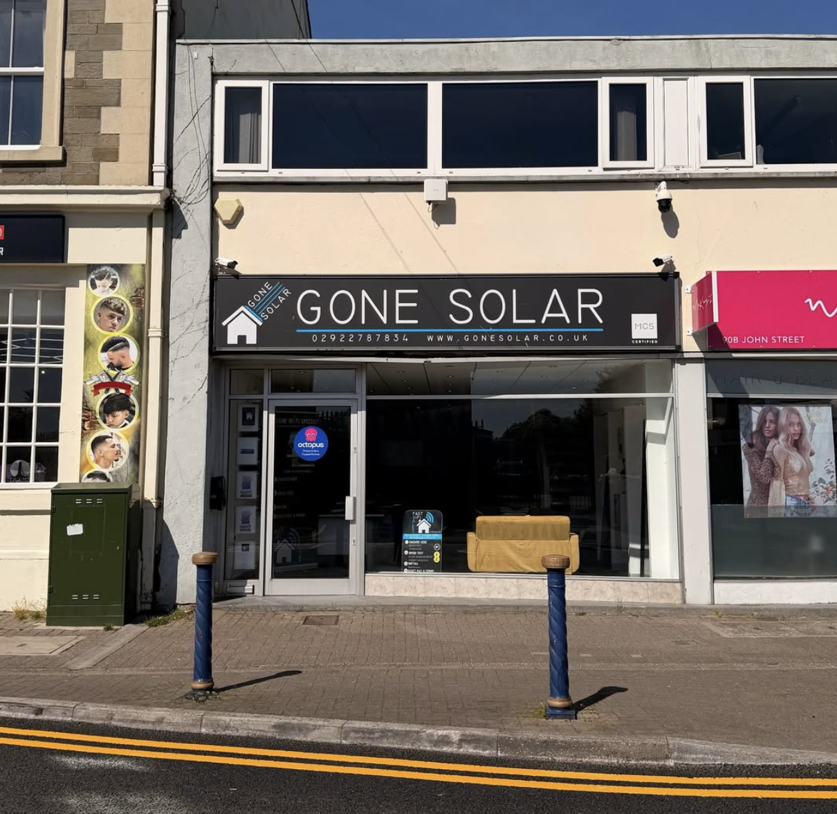 Storefront with a sign that reads "Gone Solar," featuring a house icon and contact information, with a glass door and window displaying interior furniture and signs, flanked by neighboring shop signs and posters.