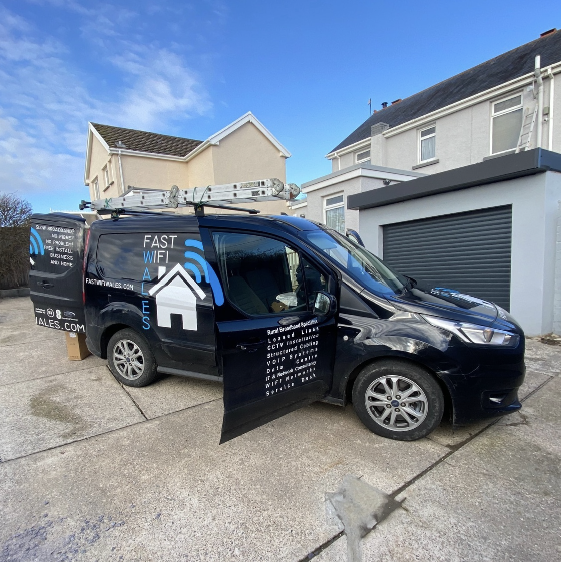 Black service van with business branding, parked on driveway near houses, with ladders on roof and open sliding door revealing company information.