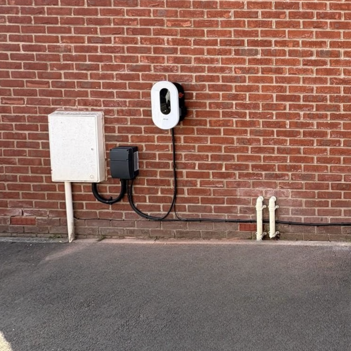Electric vehicle charging station mounted on a brick wall next to electrical boxes and pipes.