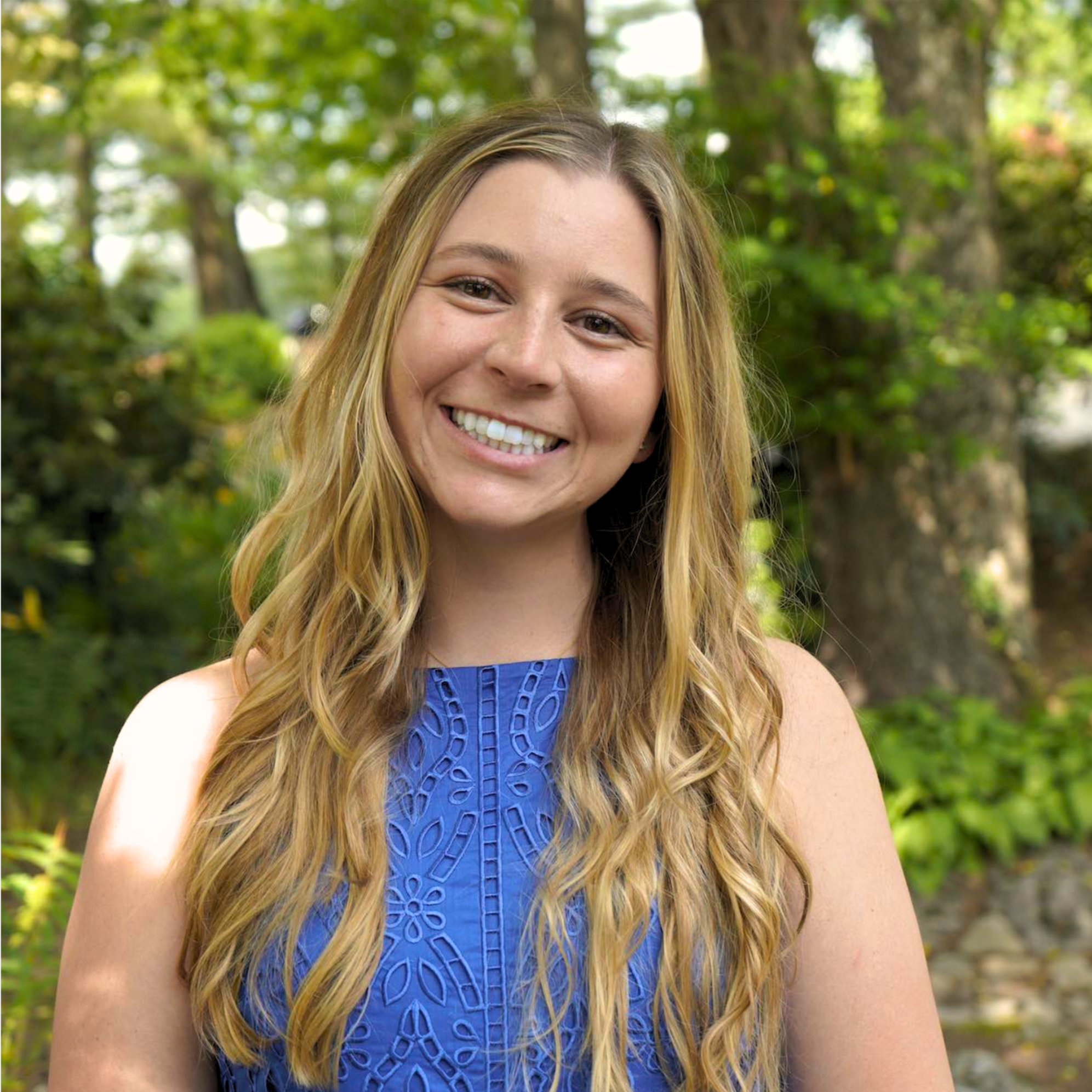 Close-up of Josi Dacko with long blonde hair wearing a blue sleeveless dress, outdoors with green trees and foliage in the background.