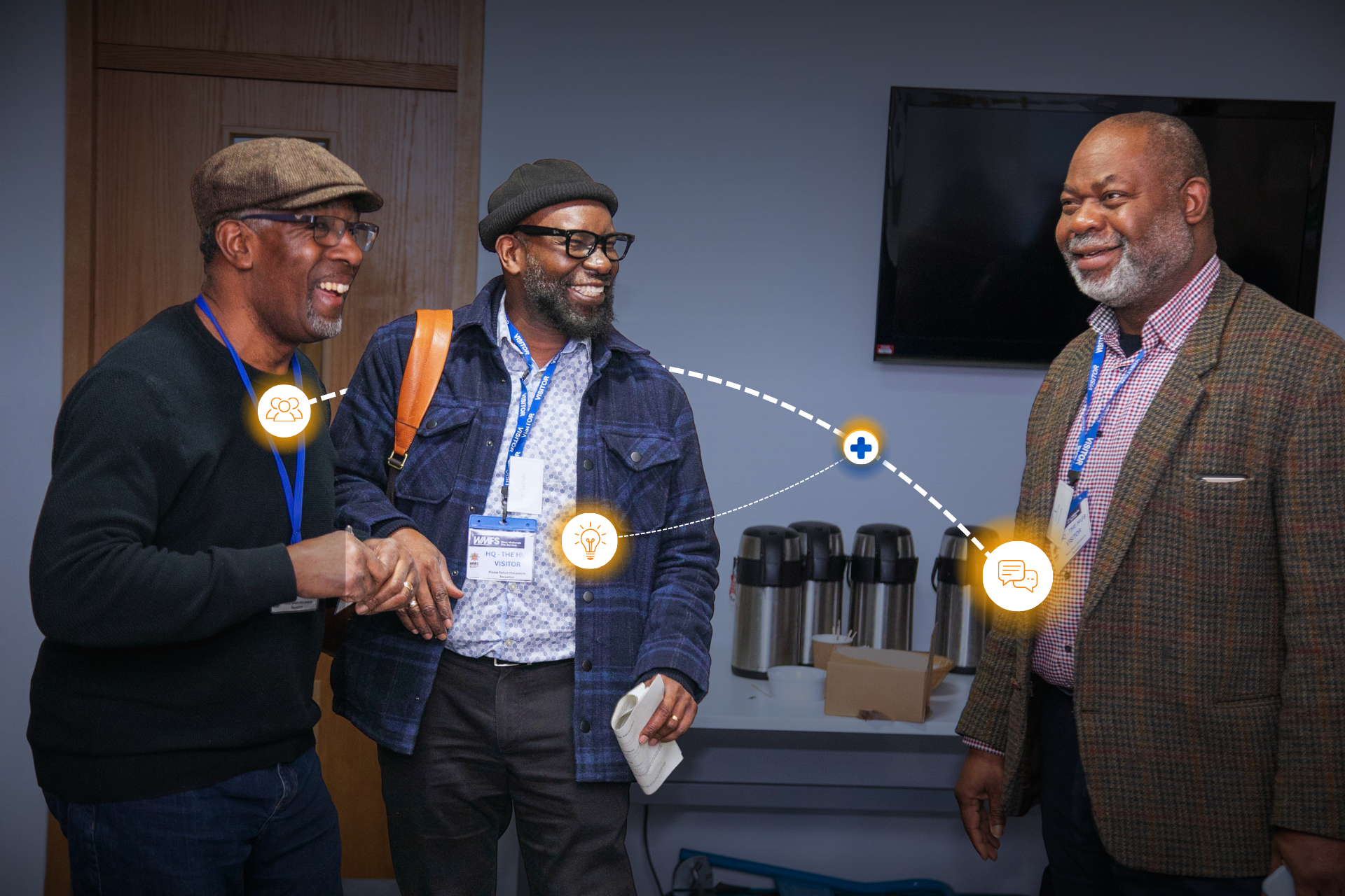 Three men in business casual attire talking and smiling in a conference room, with a television screen and coffee mugs in the background.
