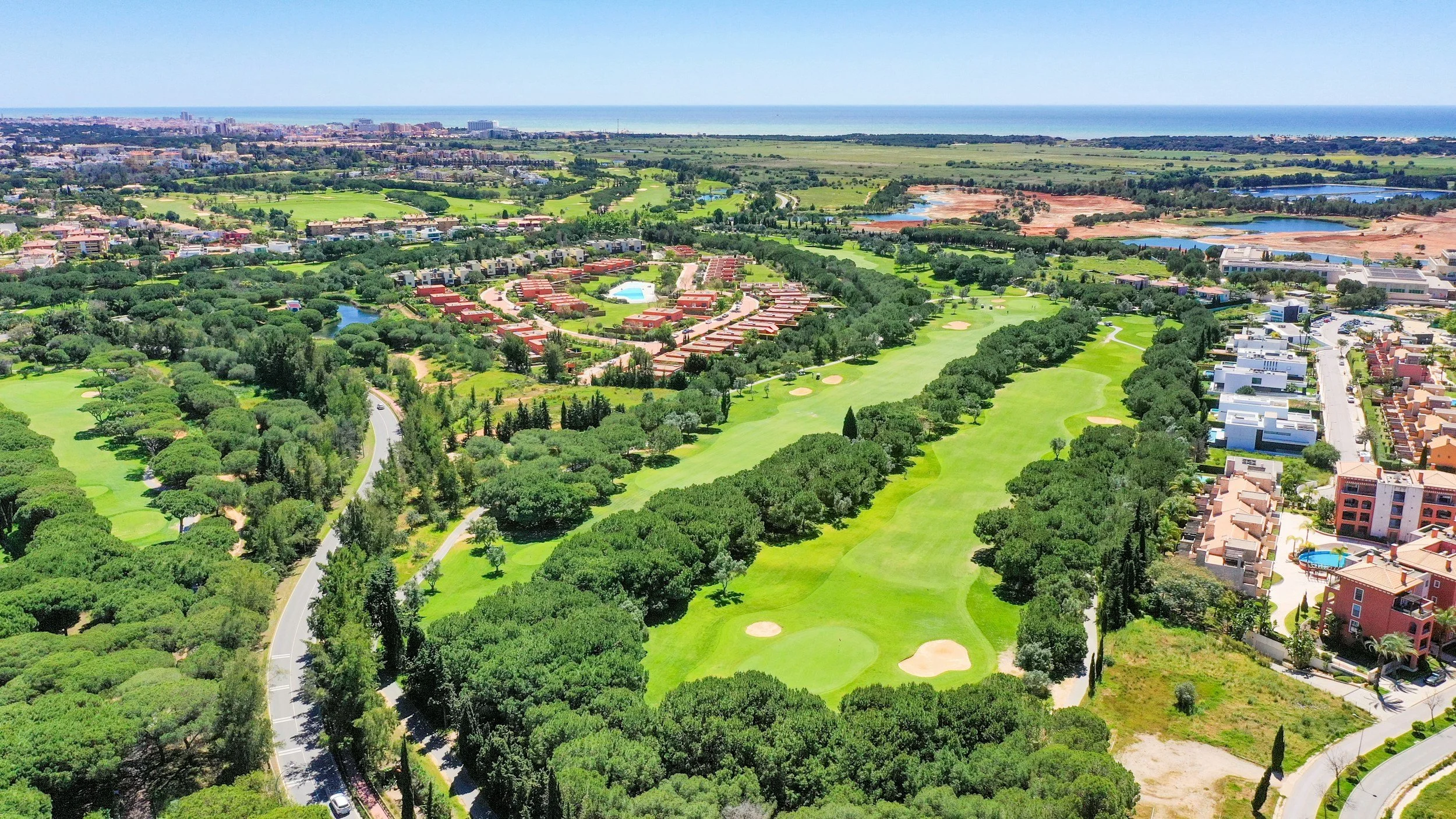 Aerial view of a golf course with green fairways, sand bunkers, surrounded by trees. Residential buildings and a cityscape are visible in the background near the coast.