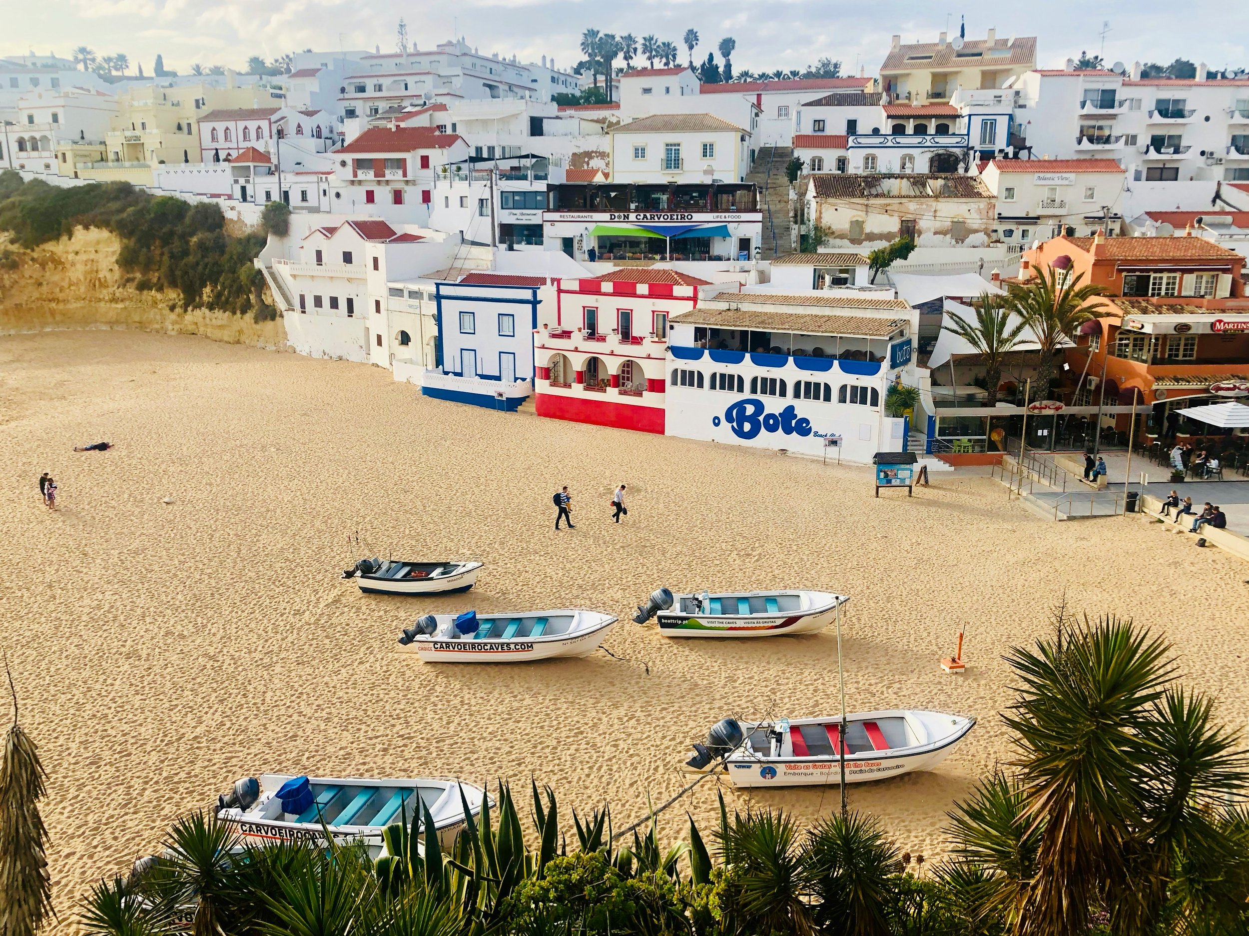 Beach with boats and white buildings on a hillside, palm trees, and people walking on the sandy shore.