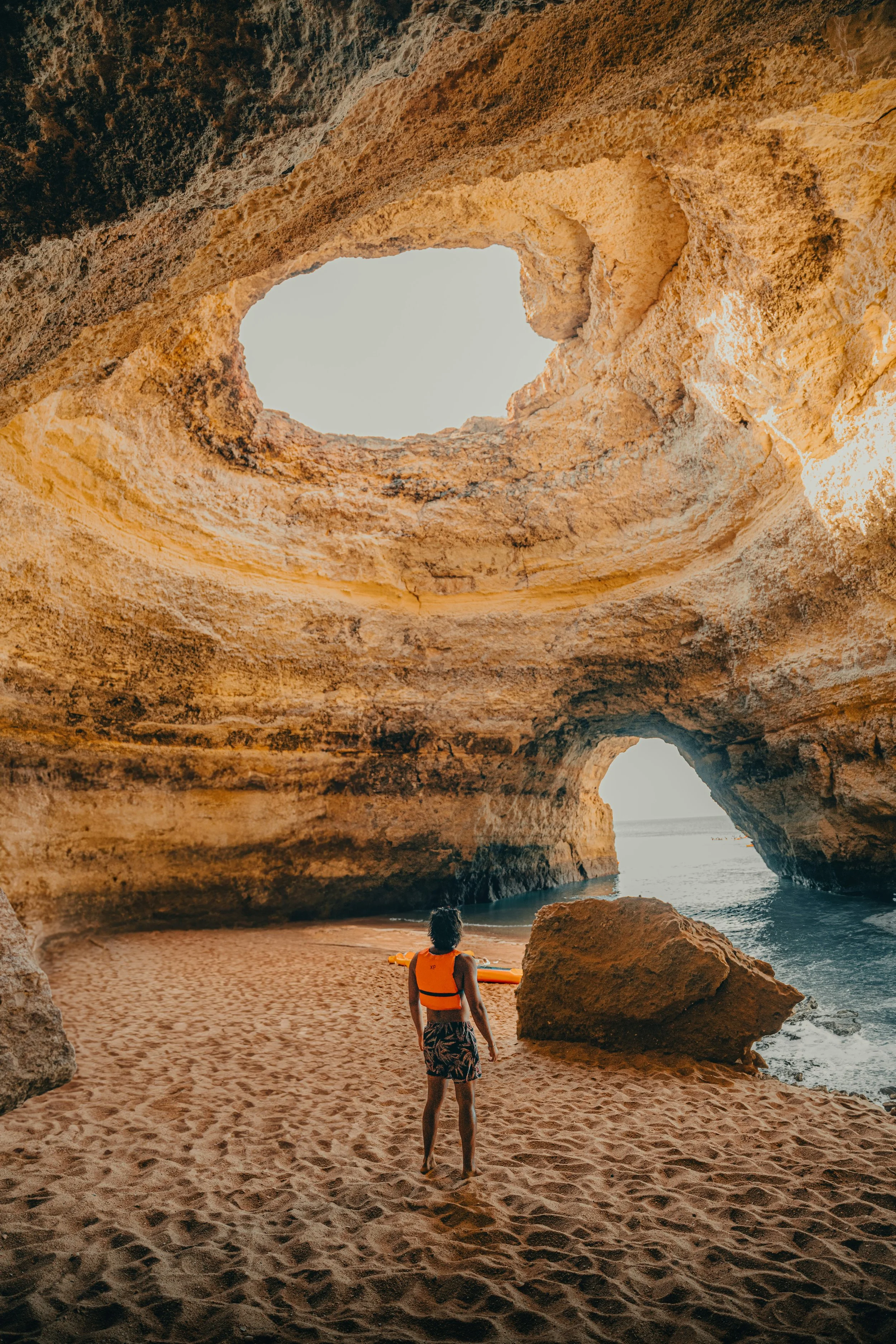 A person in a life jacket prepares to go surfing at a beach inside a large cave or rock formation with openings revealing the ocean outside.