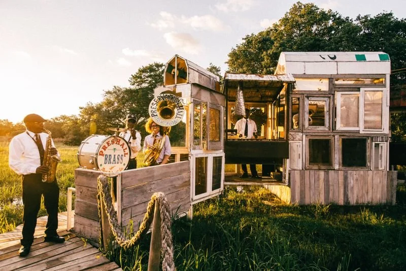 A small brass band performing outdoors on a rustic wooden stage, with one musician playing the saxophone, a bass drum, and a tuba, surrounded by grassy fields and trees during sunset.
