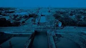 A cityscape view featuring a large bridge crossing over a water body, with buildings and structures in the background under a dark sky.