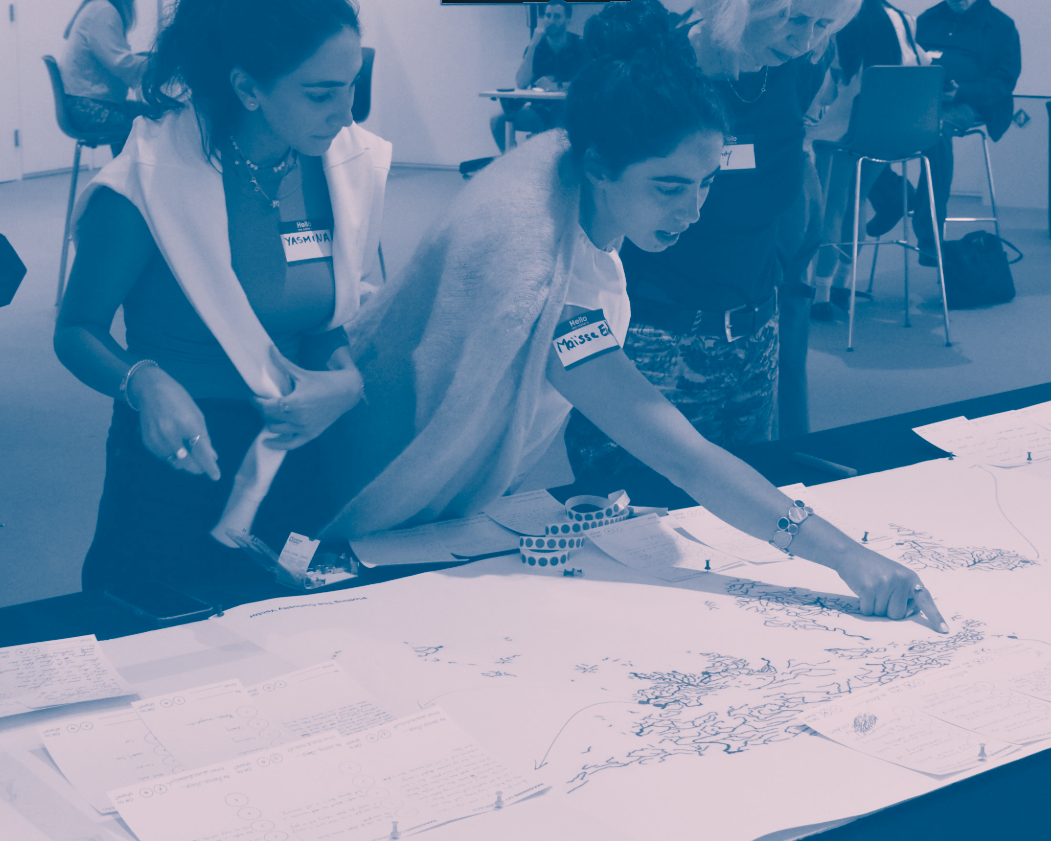 Two women and one person working together at a table, examining and pointing at large sheets of paper with handwritten notes and diagrams in a classroom or workshop setting.