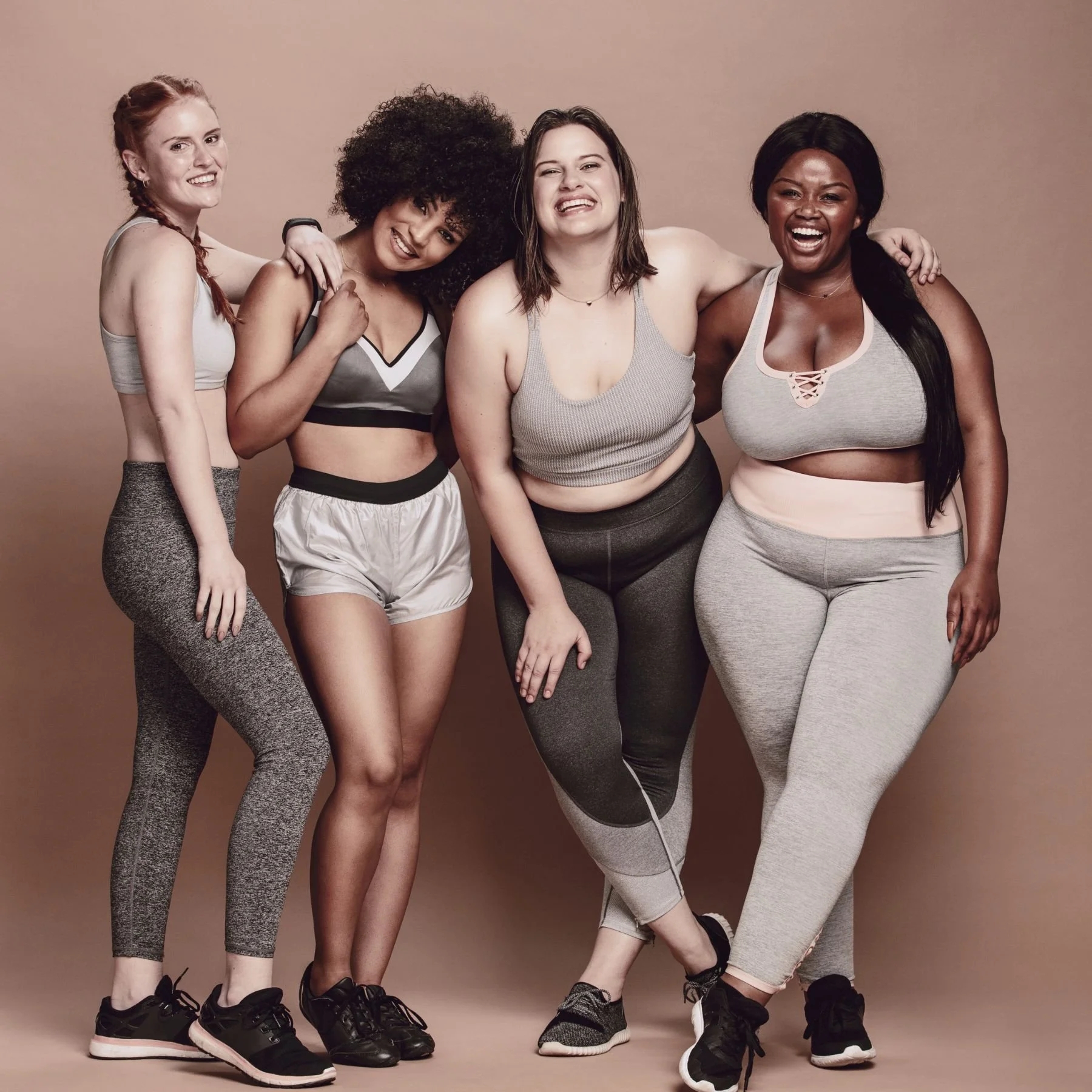 A diverse group of five women in athletic wear standing together against a neutral background, smiling and laughing, with their arms around each other.
