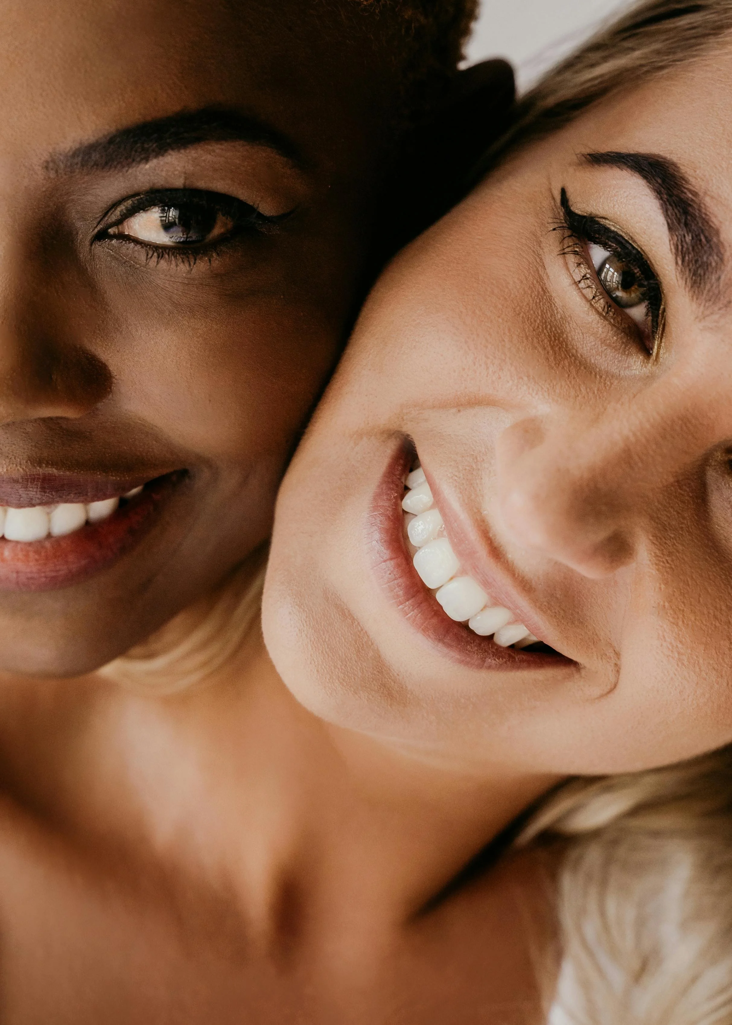 Close-up of two smiling women, one with dark skin and the other with light skin, showing their eyes, eyebrows, and teeth, with their heads close together.