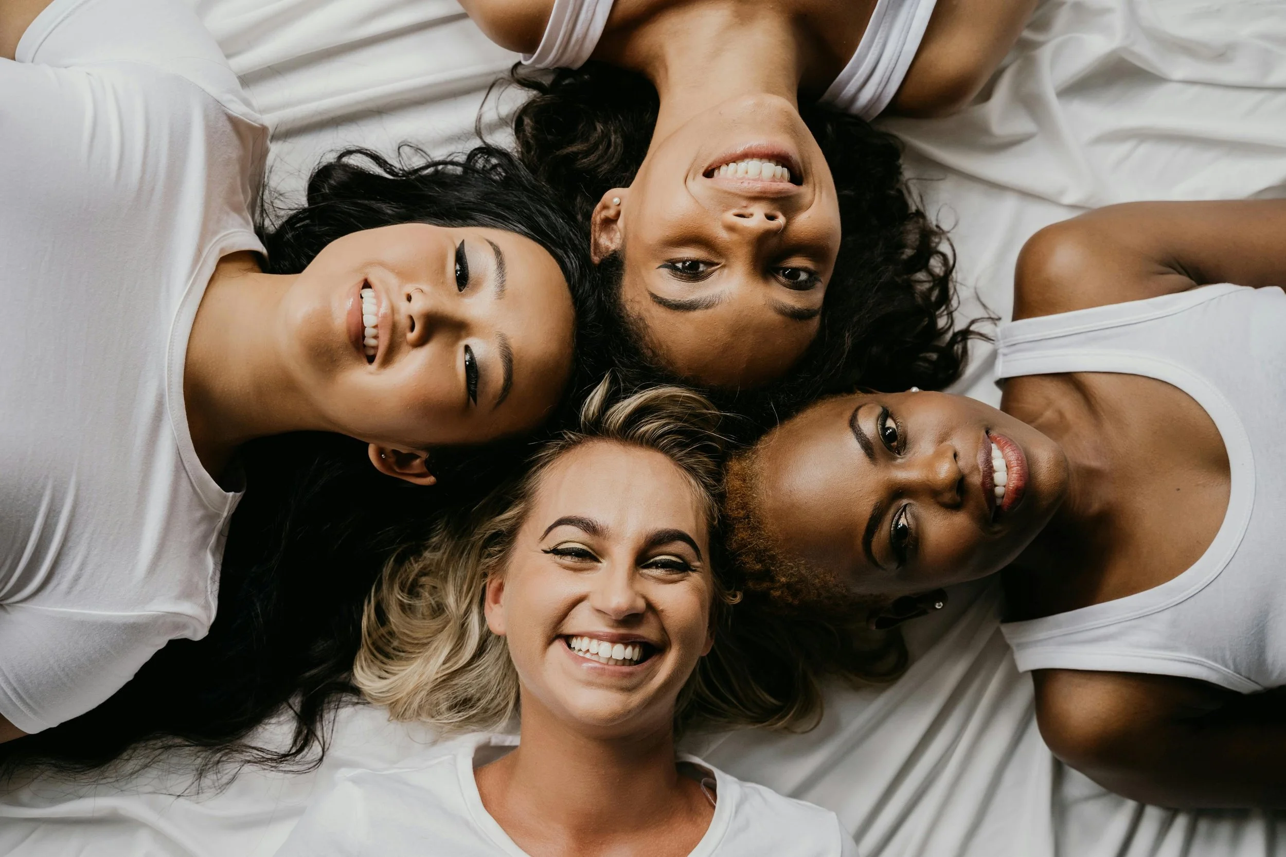 Four women of diverse ethnicities lying on a white surface, their heads close together, smiling and looking at the camera.