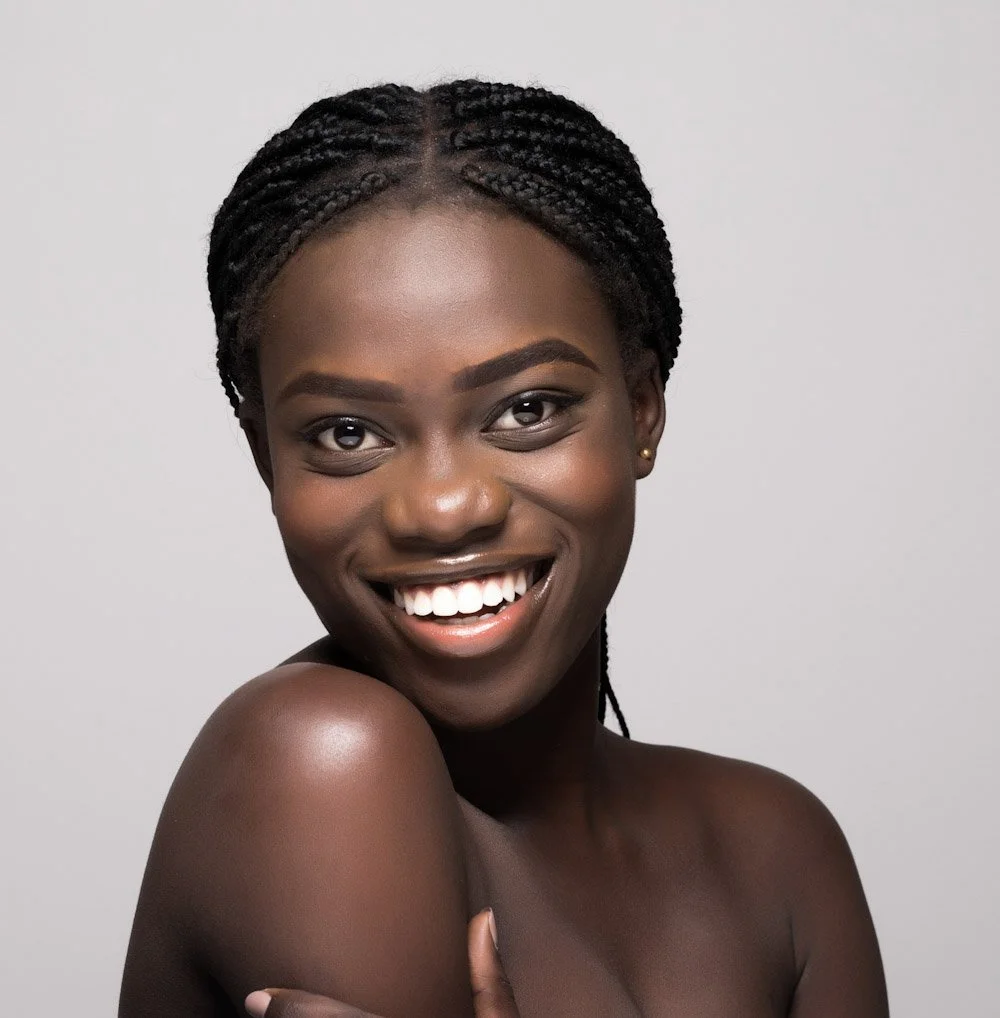 A smiling young Black woman with braided hair and minimal makeup, posing against a plain gray background.