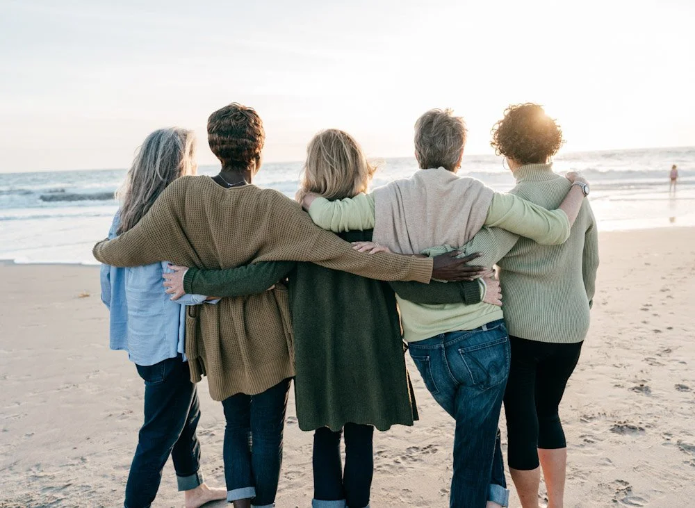Five women standing on a beach, facing the ocean, with their arms around each other during sunset.