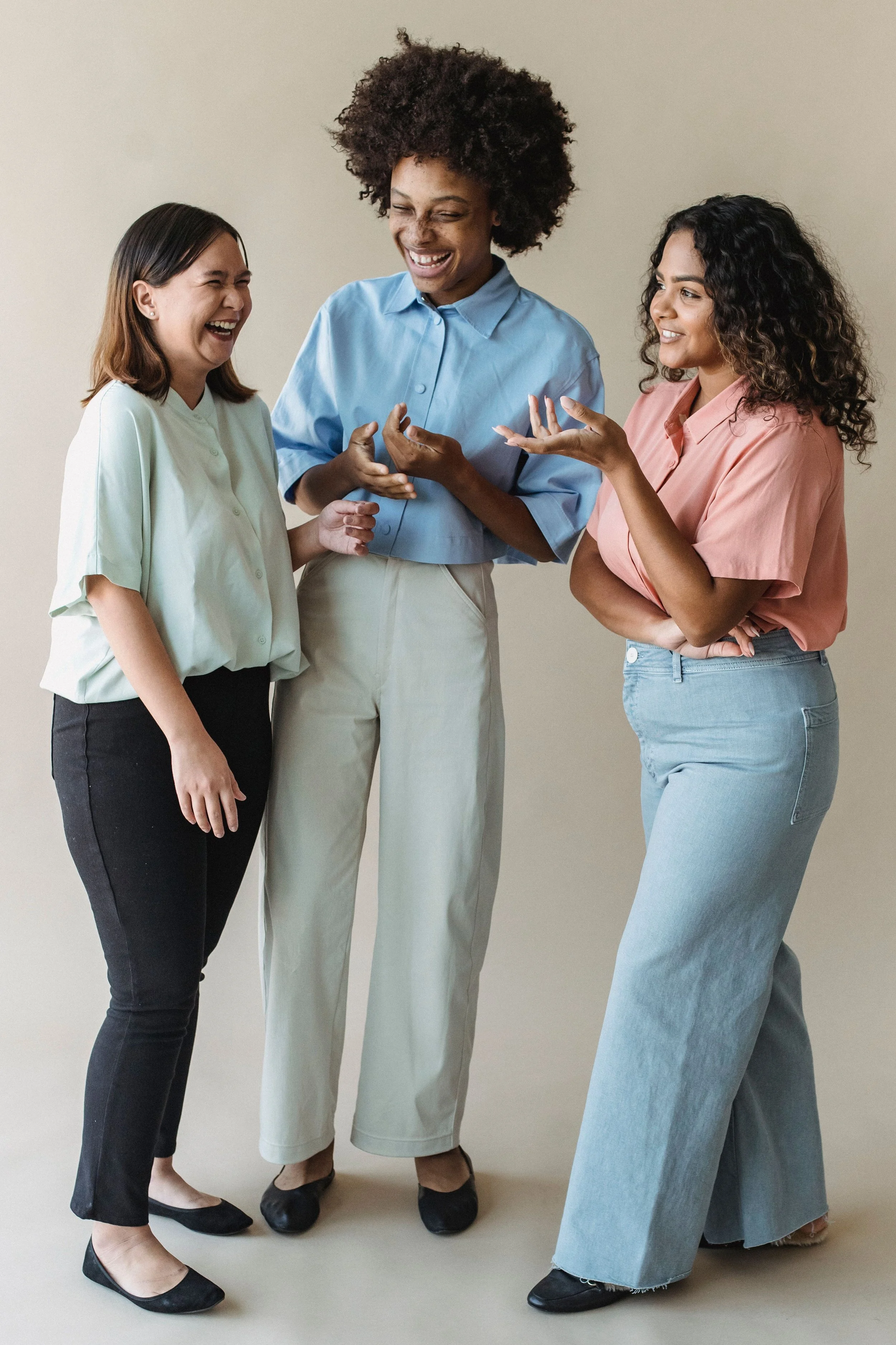 Three diverse women standing together, laughing, and conversing in an indoor setting with a plain background.