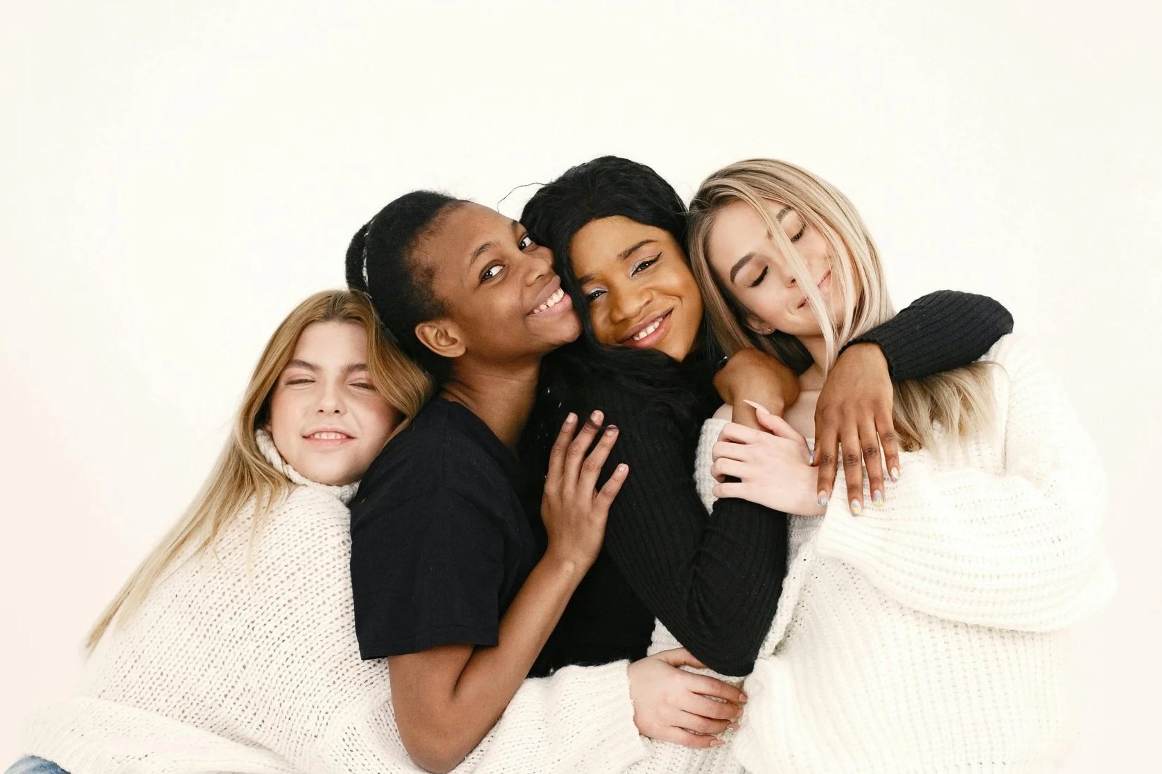 Four women hugging and smiling, standing closely together against a plain white background. They appear happy and affectionate.