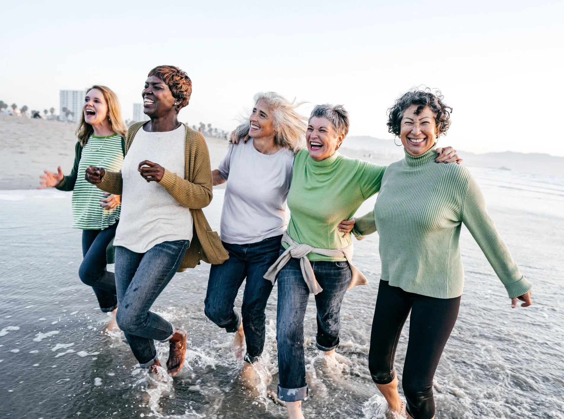 Group of five women enjoying a walk along the beach shoreline with ocean waves and buildings in the background.