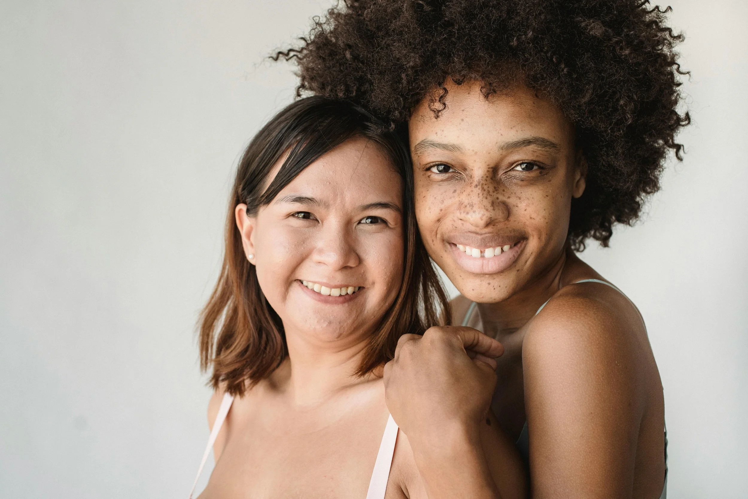 Close-up of two women embracing and smiling at the camera, one with curly hair and the other with straight hair, against a plain, light-colored background.