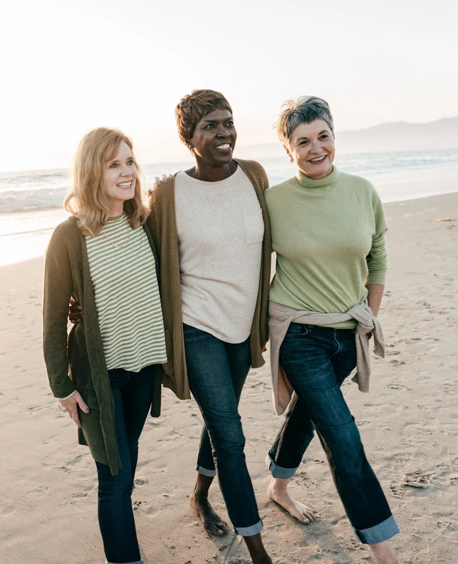 Three women walking barefoot on the beach, smiling and enjoying each other's company during sunset.