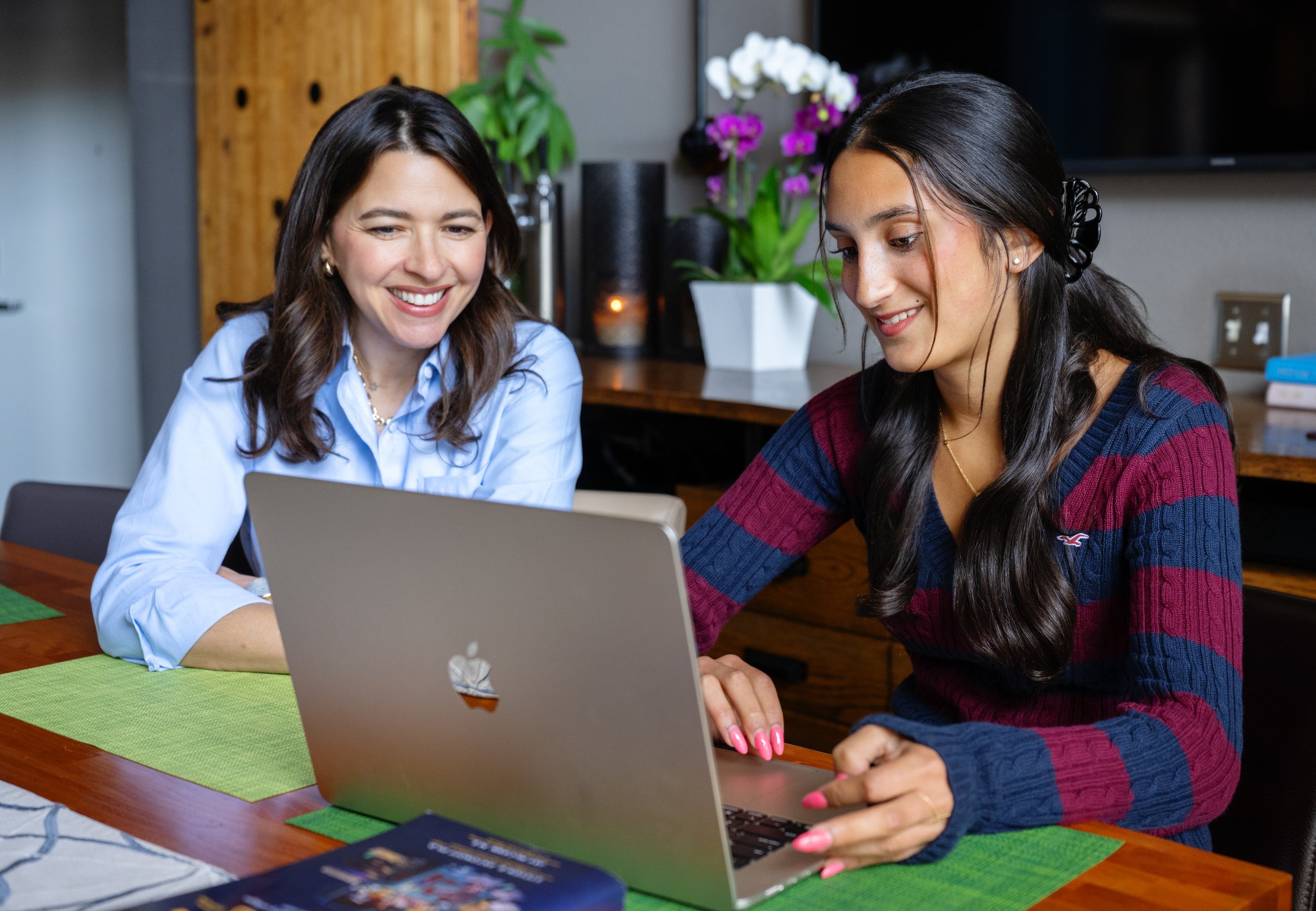 Two women are sitting at a wooden table looking at a laptop screen together, smiling. One woman has long dark hair, wears a light blue blouse, and the other has dark hair tied back with a black hair clip and is wearing a red and blue striped sweater. Behind them, there are potted plants with purple and white flowers, and a wooden cabinet with a candle or small lamp on it.
