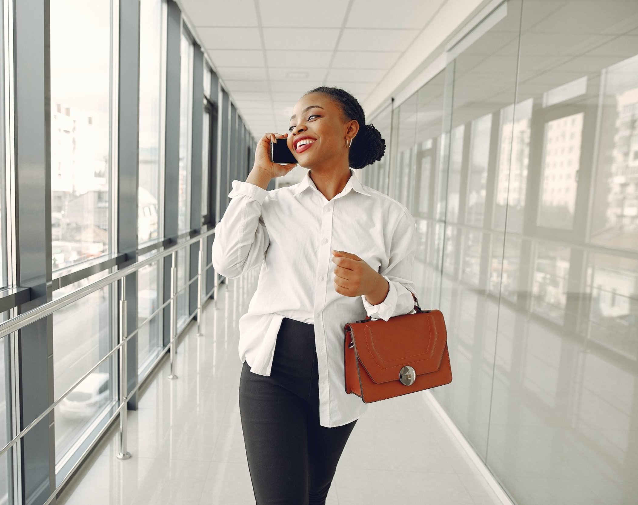 A woman with braided hair, wearing a white shirt and black pants, is talking on her phone while standing in a modern building with large windows and a city view, holding a brown handbag.