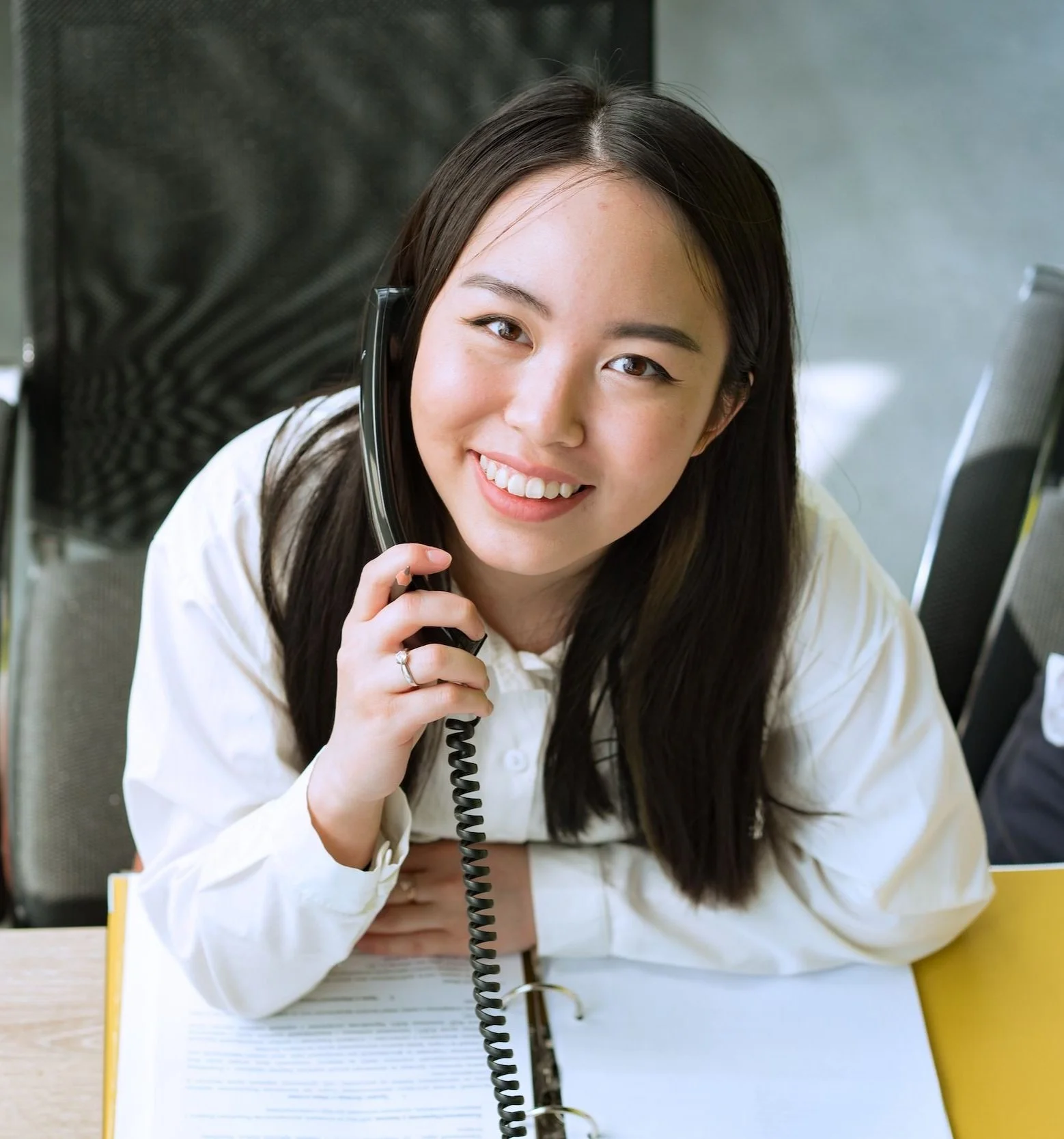 A woman with long brown hair smiling, holding a corded black telephone receiver to her ear. She is seated at a table with open notebooks and a yellow folder in front of her. The background shows office chairs.