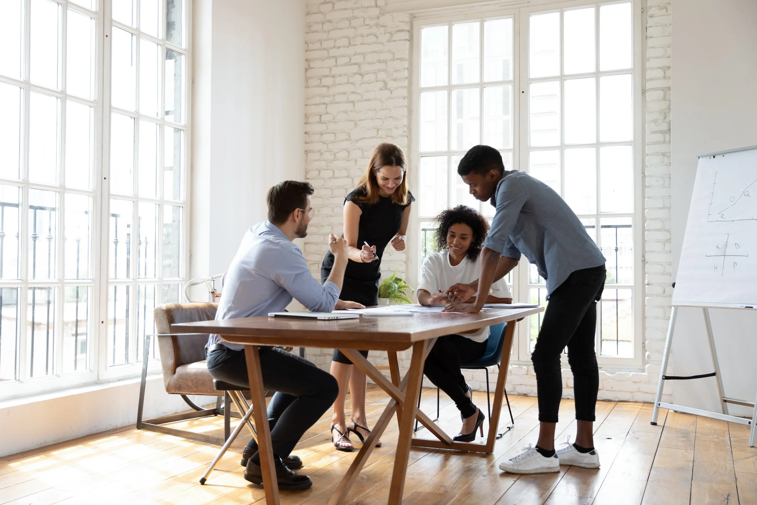 A group of four diverse people collaborate around a table in a bright, modern office with large windows and white brick walls.